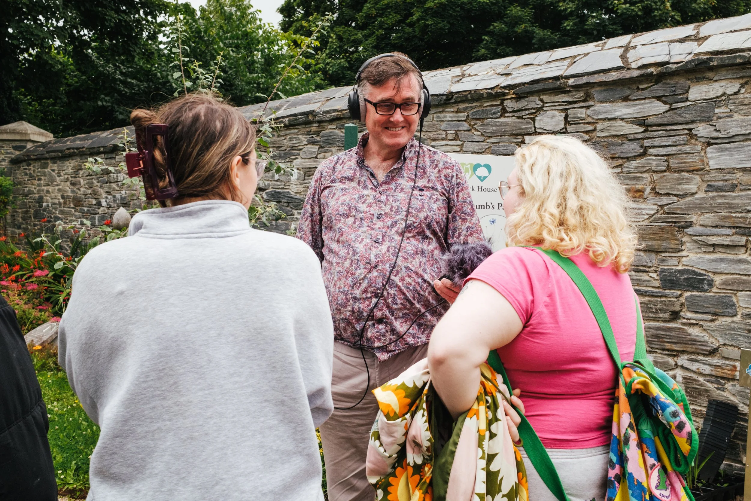 Three women and a man standing outdoors near a stone wall, engaged in conversation. The man wears glasses and headphones, and the women are dressed casually, one with a floral-patterned backpack. There are flowers and greenery around.