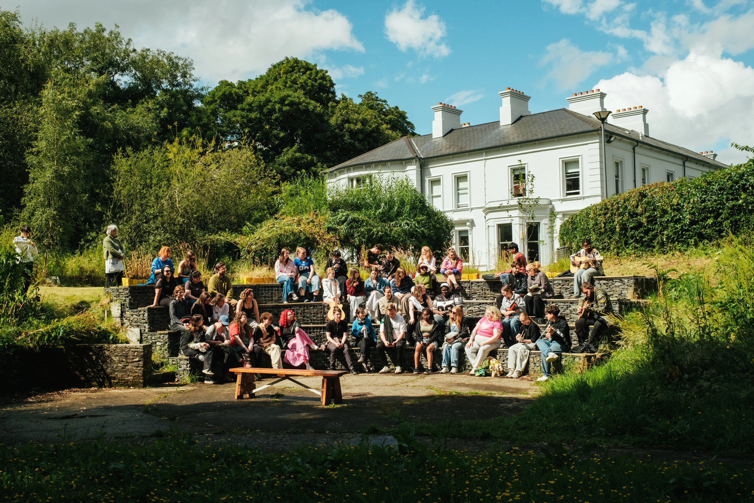 A large gathering of people sitting on a tiered outdoor amphitheater area with green trees and a white house in the background, some playing musical instruments.