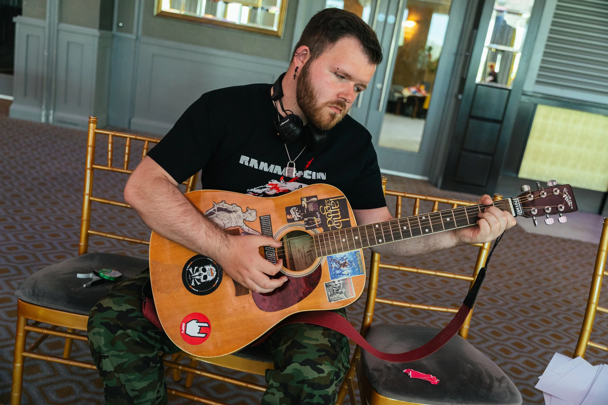 A man with short dark hair, a beard, and a pierced eyebrow sits on a gold-colored chair, playing an acoustic guitar adorned with various stickers. He is wearing a black Ramstein T-shirt, camouflage pants, and has black headphones draped around his ne