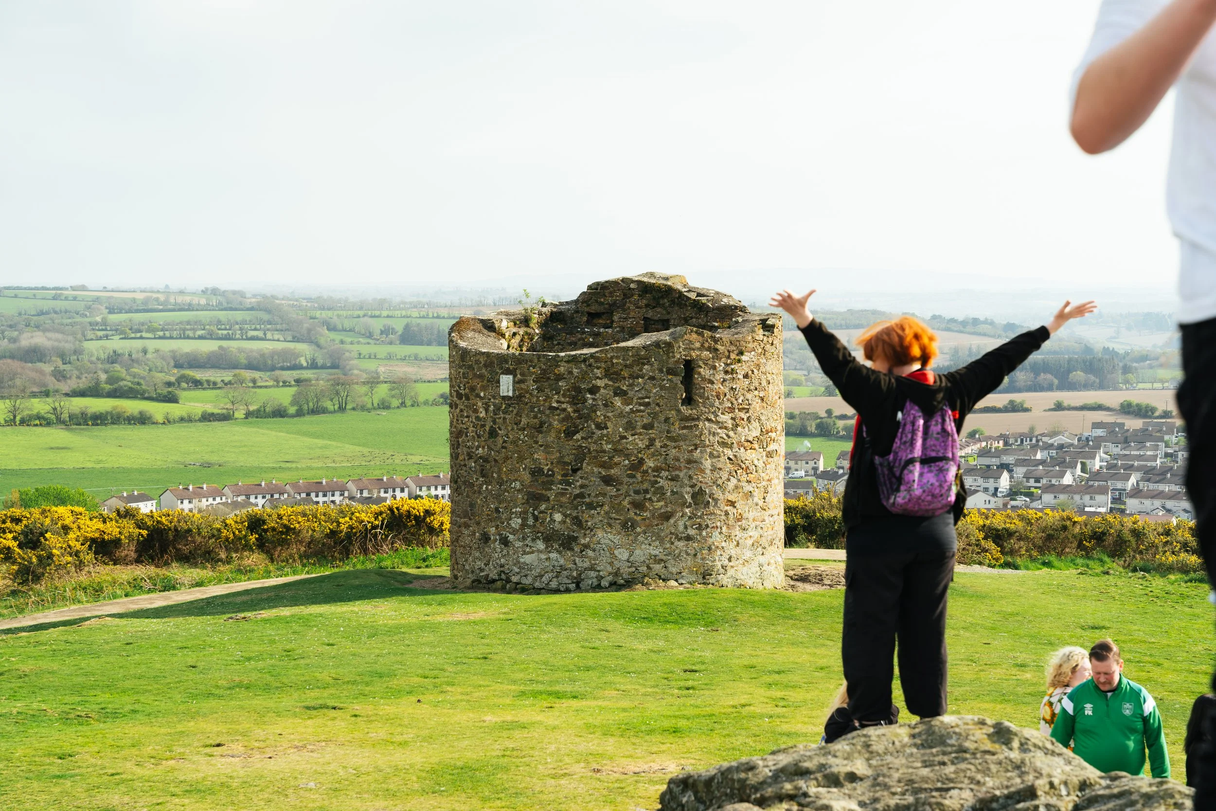 A person with orange hair and a purple backpack stands on a grassy hill with arms raised, facing an ancient stone tower with a rural landscape in the background.