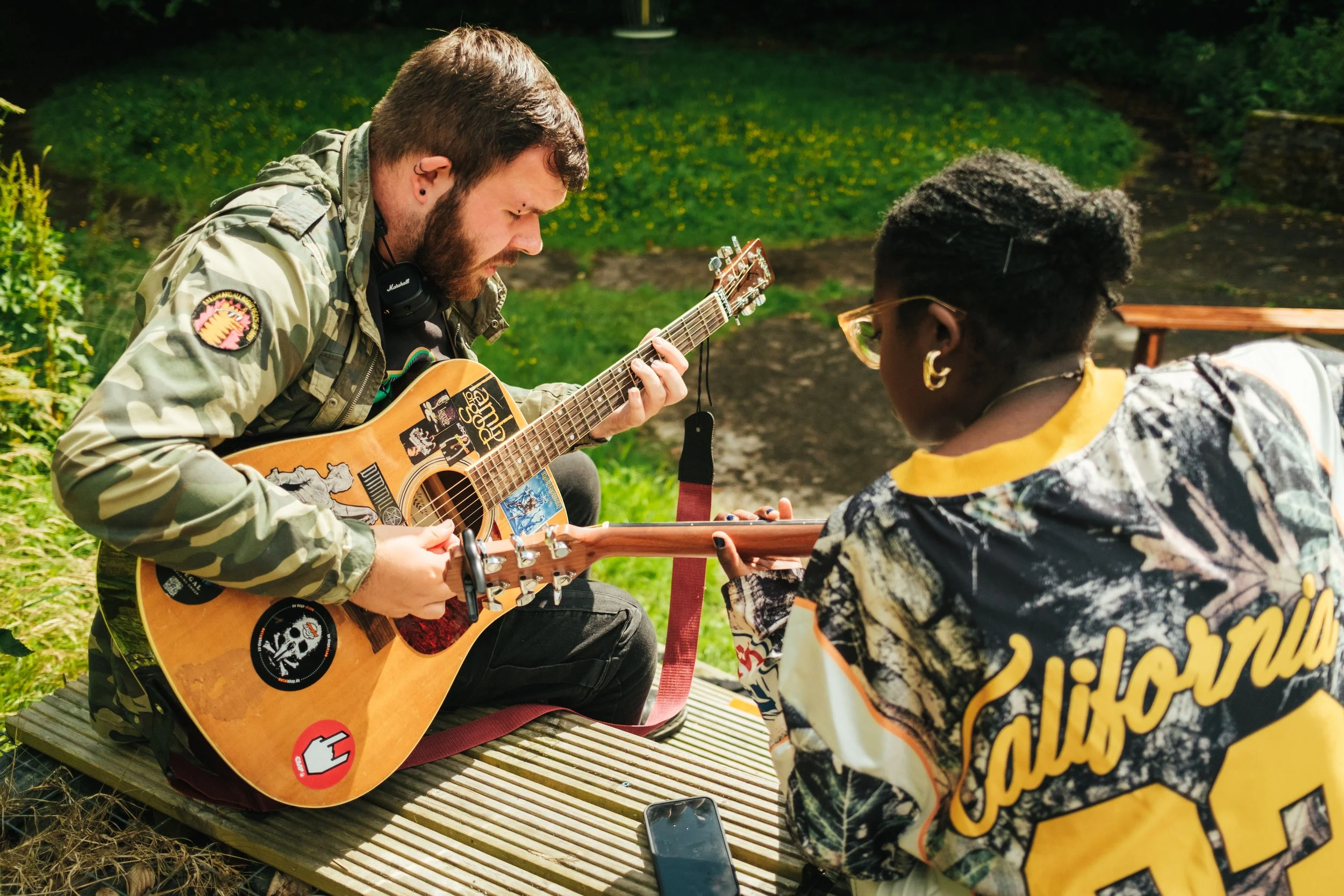 Two people sitting outdoors on a wooden platform, playing guitars and gazing at each other. One is wearing a camouflage jacket and headphones, the other has short hair, glasses, and a jacket with 'California' written on the back.