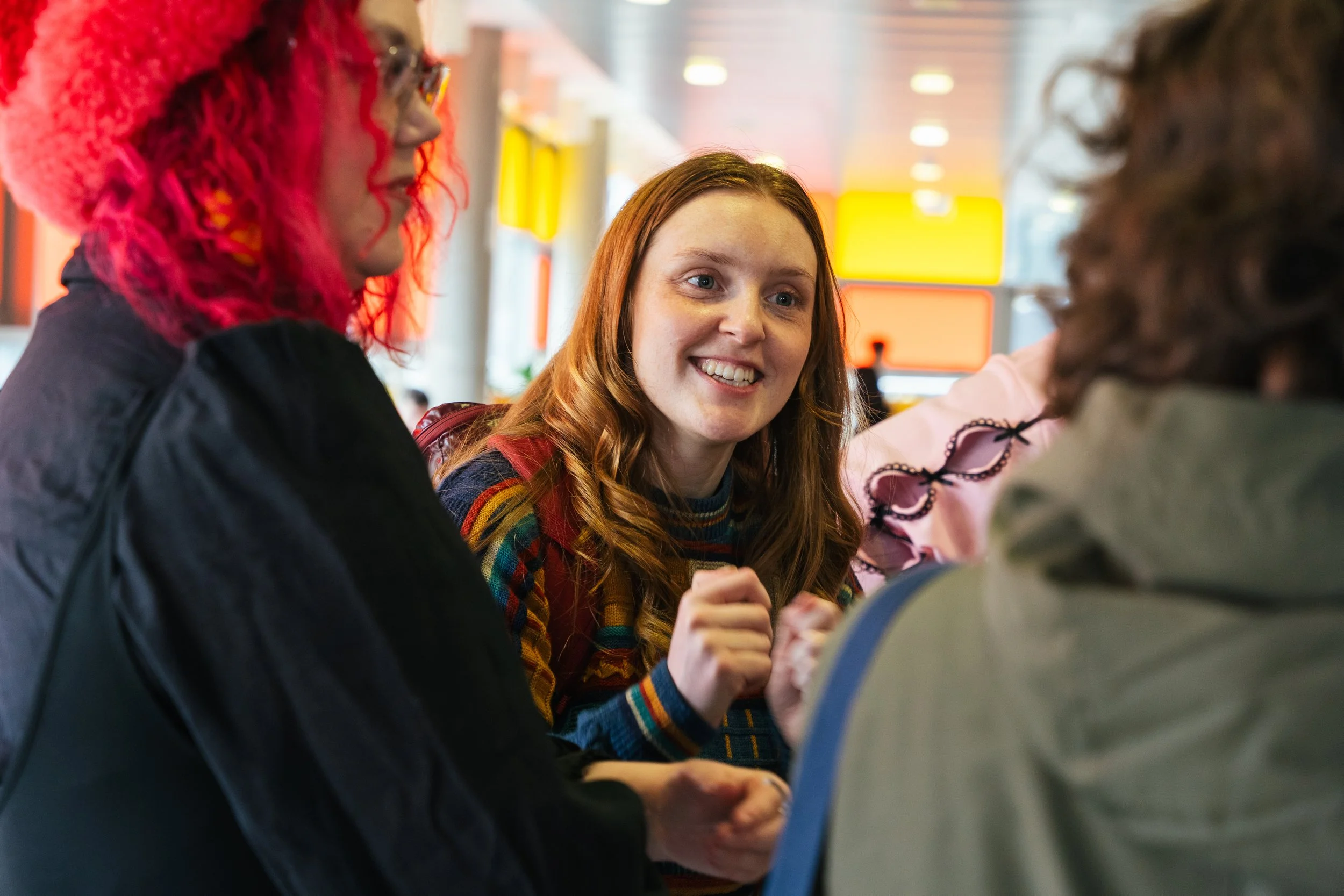 A group of three young women sitting together and talking in a brightly lit indoor space, with one woman smiling and making eye contact with the others.