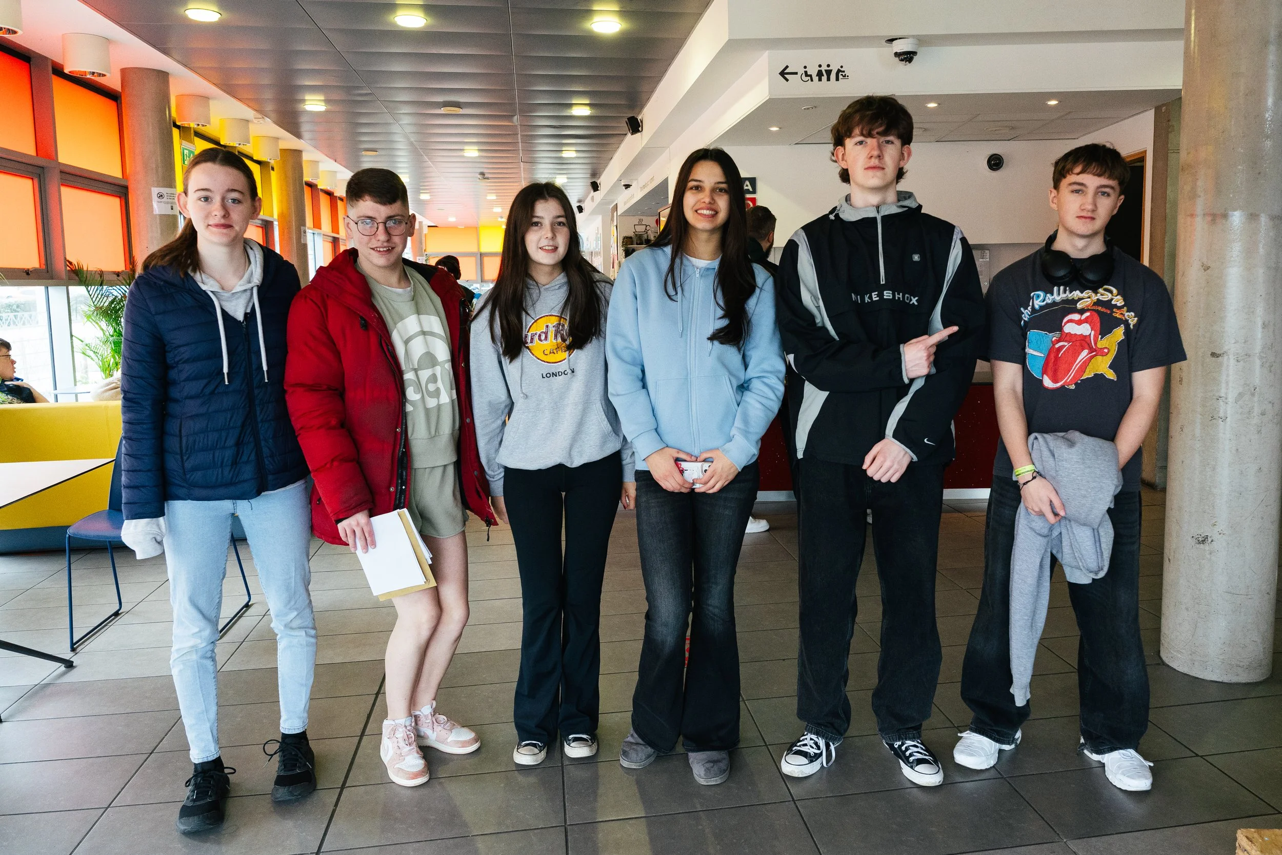 Six teenagers standing in a row inside a modern building. They are dressed casually with jackets, hoodies, and sneakers. Some are holding notebooks or jackets. The background shows colorful window panels and a seating area.