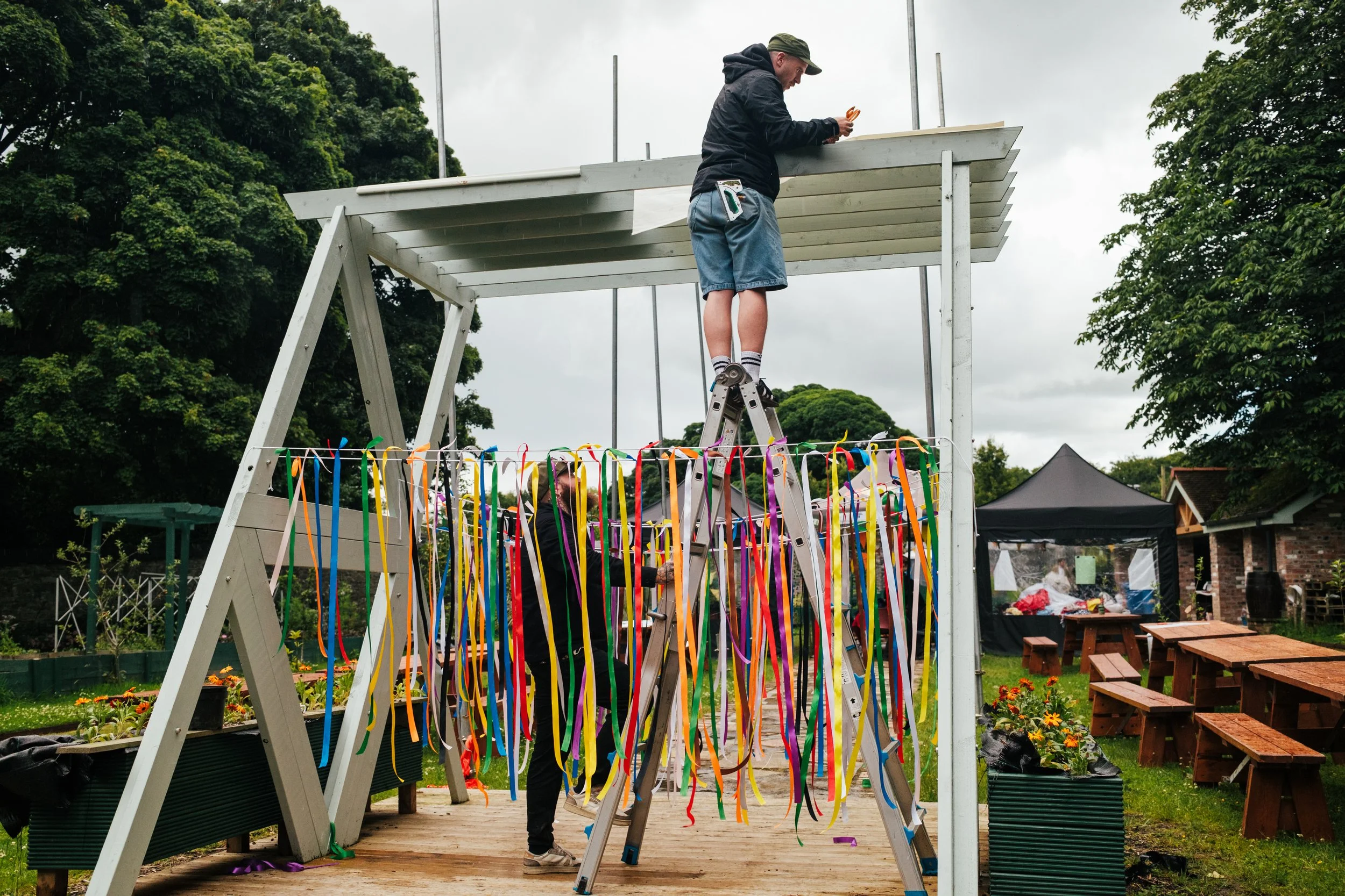 Two people are working on a wooden structure outdoors on a cloudy day. One person is standing on a ladder, working on the roof, while another person is below, handling colorful ribbons that are hanging from the structure. In the background, there are