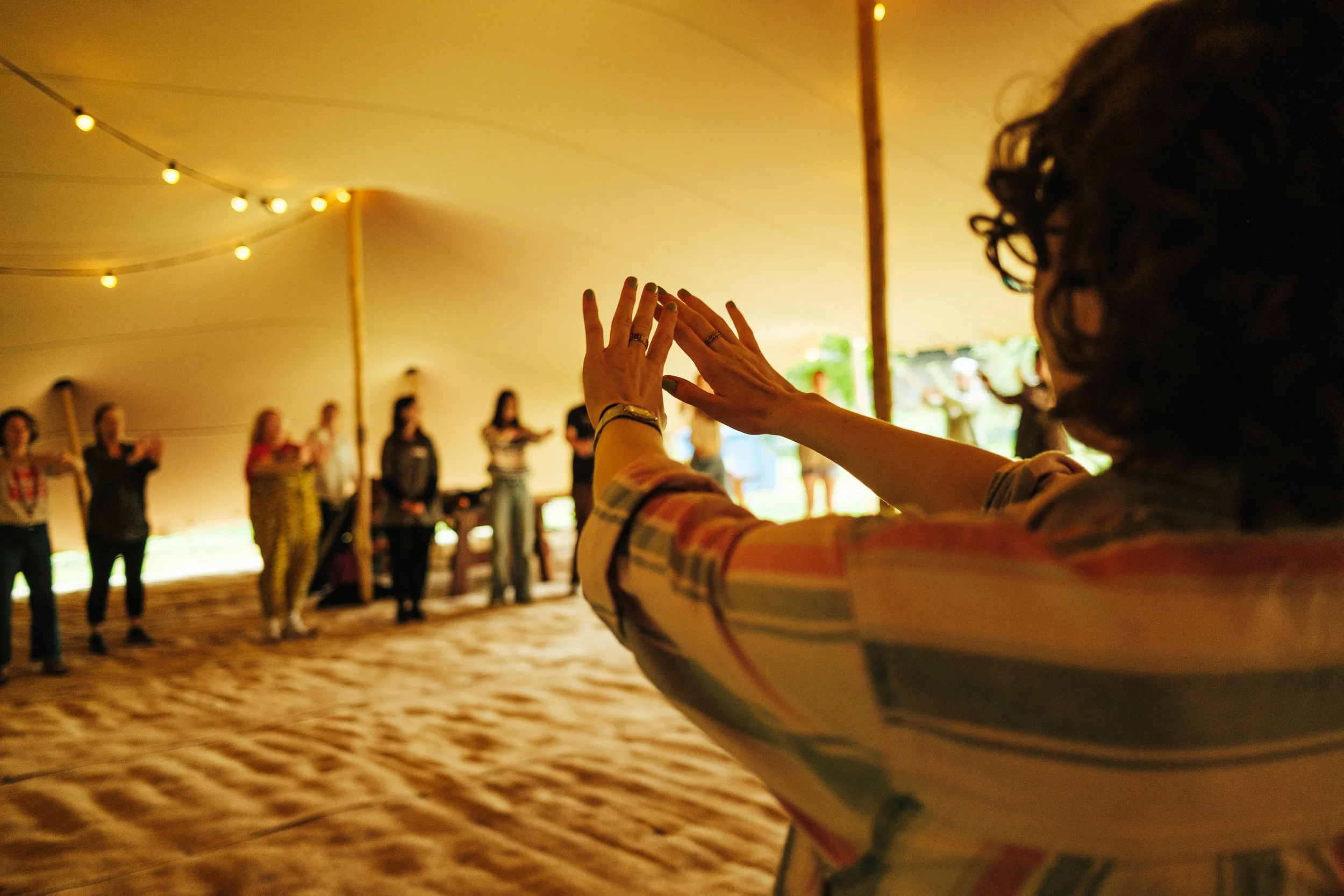 A woman with curly hair and a plaid shirt stands with her arms raised in front of a group of people under a large tent at night. The group is standing in a semi-circle, with some raising their hands. String lights are hanging from the ceiling of the 