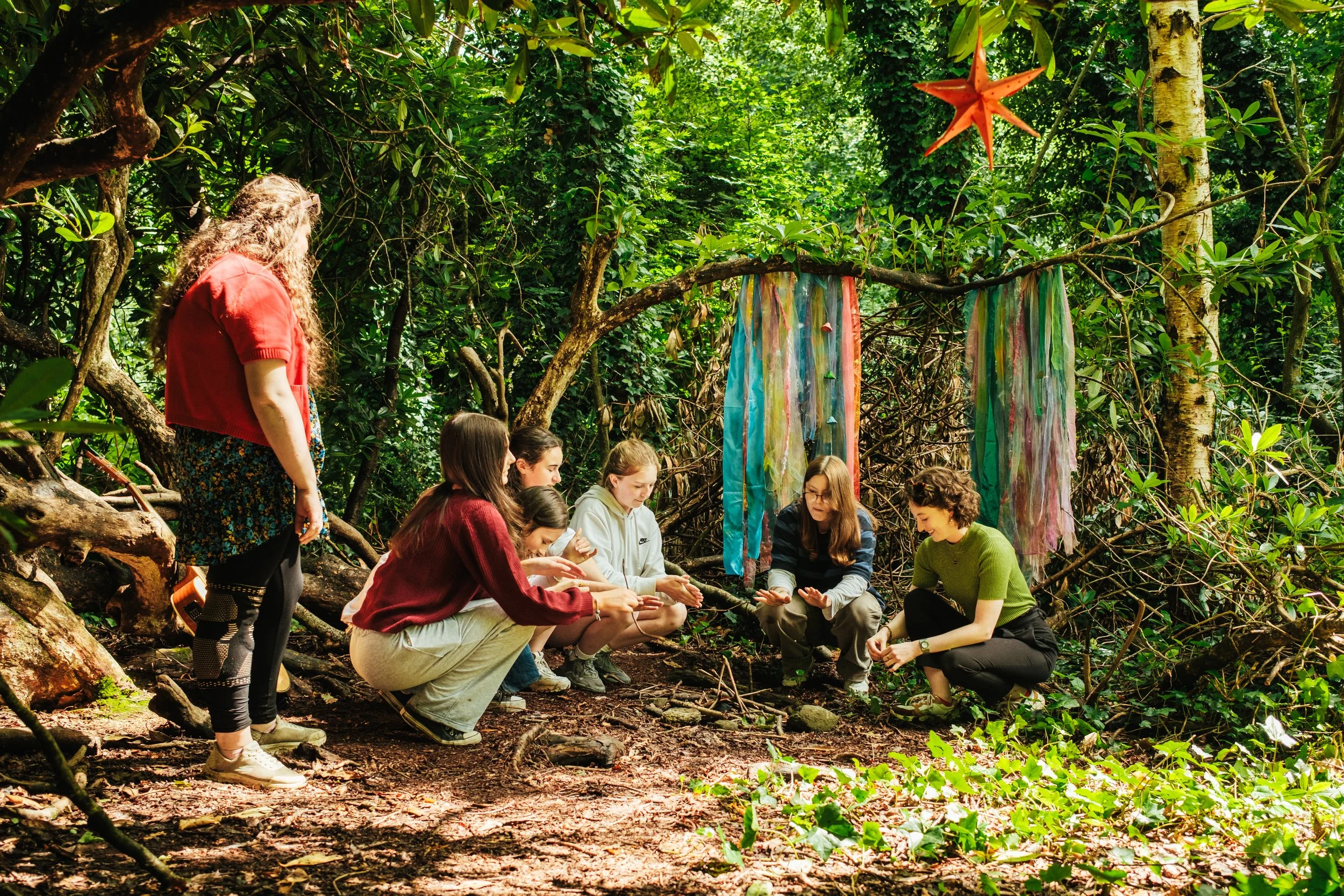 Group of children and a woman sitting and kneeling in a forest, engaging in an outdoor activity, with colorful fabric decorations hanging in the background.