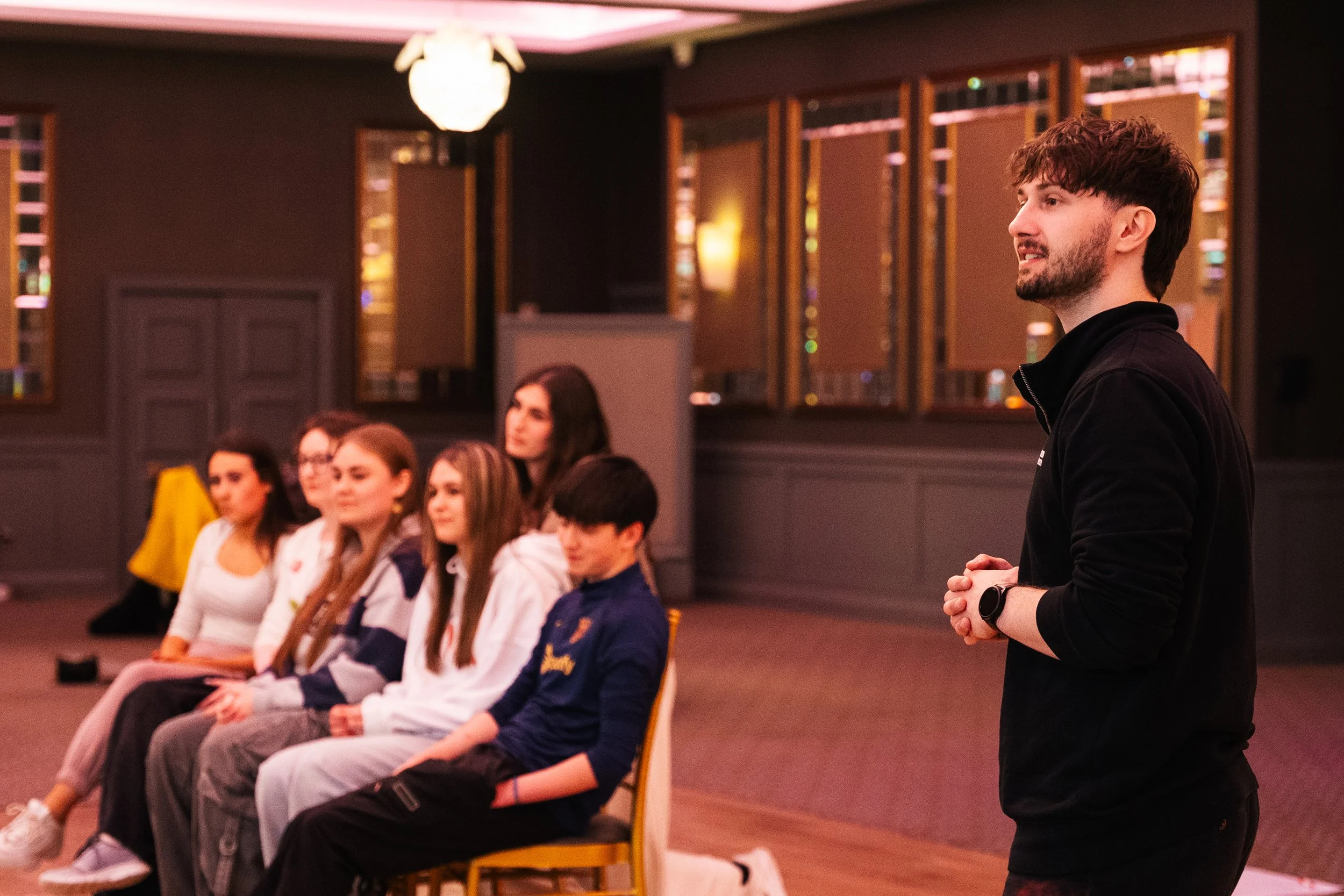 A man giving a presentation to a group of young women and one young man seated in chairs in a dimly lit room with mirrors and warm lighting.