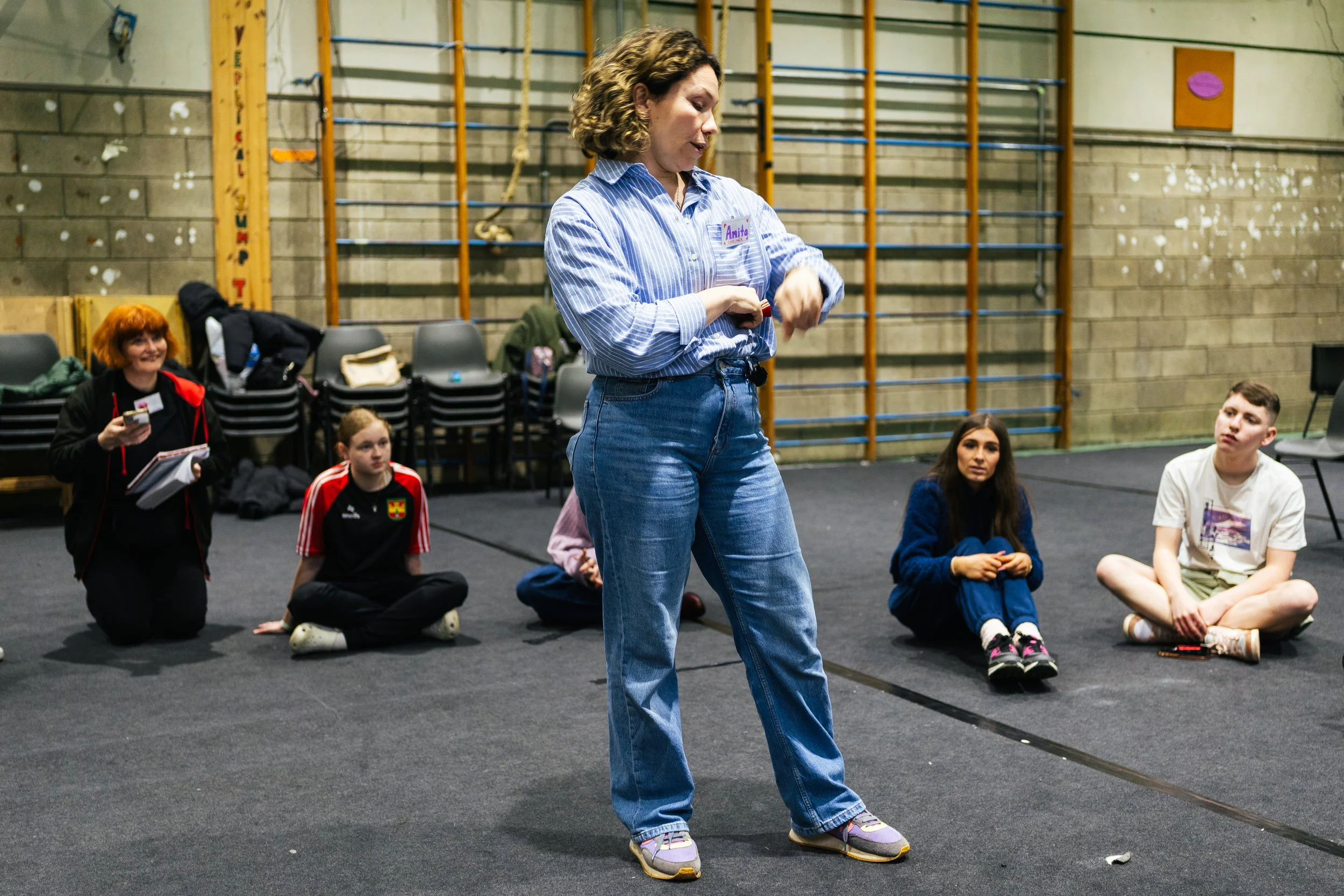 A woman with curly hair wearing a blue and white striped shirt and jeans, standing and talking to a group of young people sitting on the floor in a gymnasium.