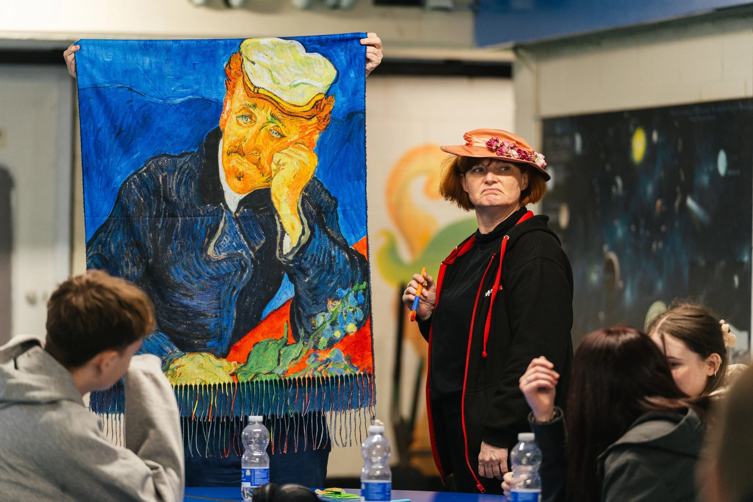 A woman with a pink hat decorated with flowers holds a marker, standing next to a tapestry of Vincent van Gogh's painting 'The Potato Eaters' displayed at a gathering of young people sitting at a table