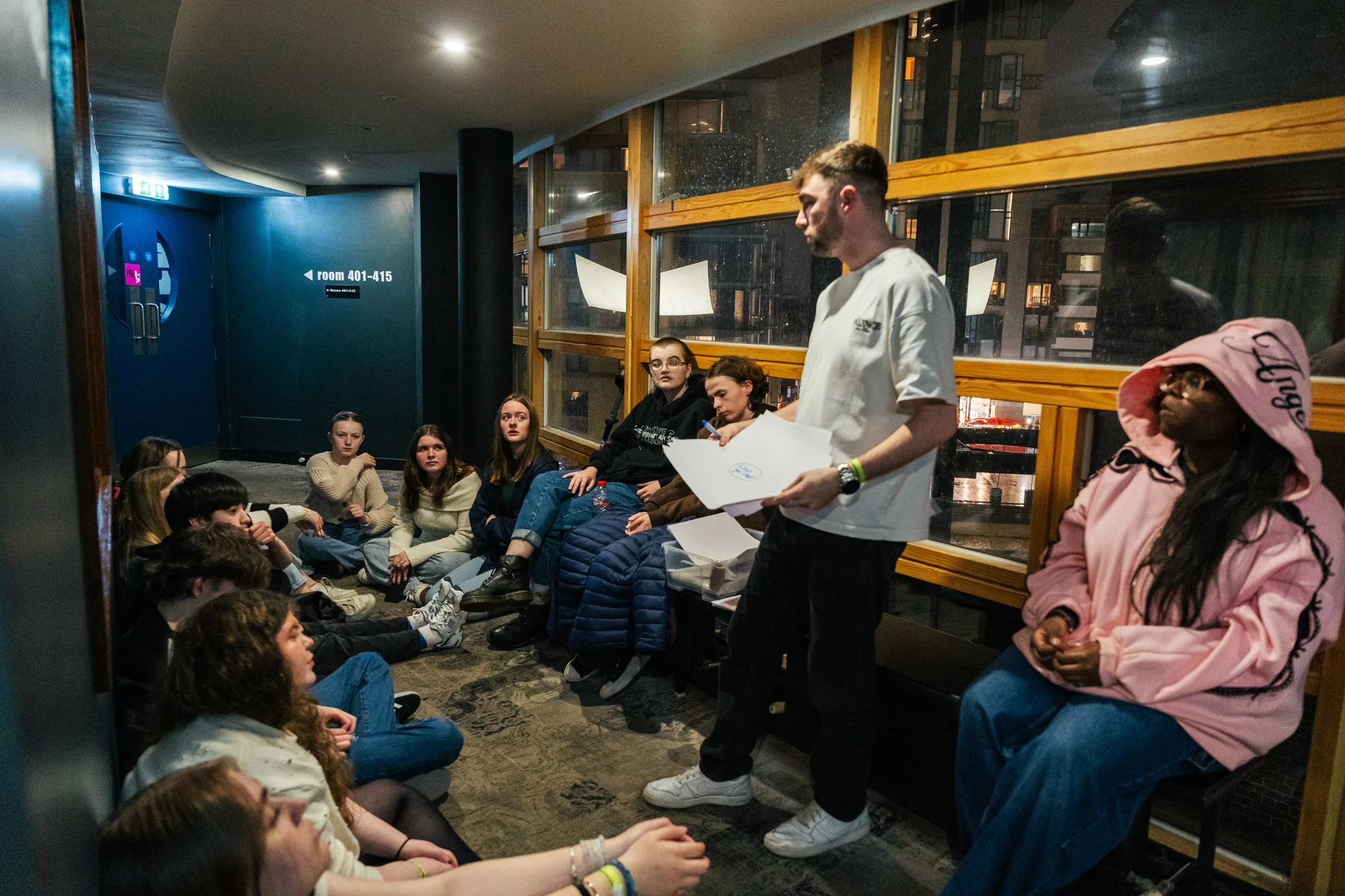A group of young people texting a man standing and speaking in a dimly lit room with large windows and wooden accents.
