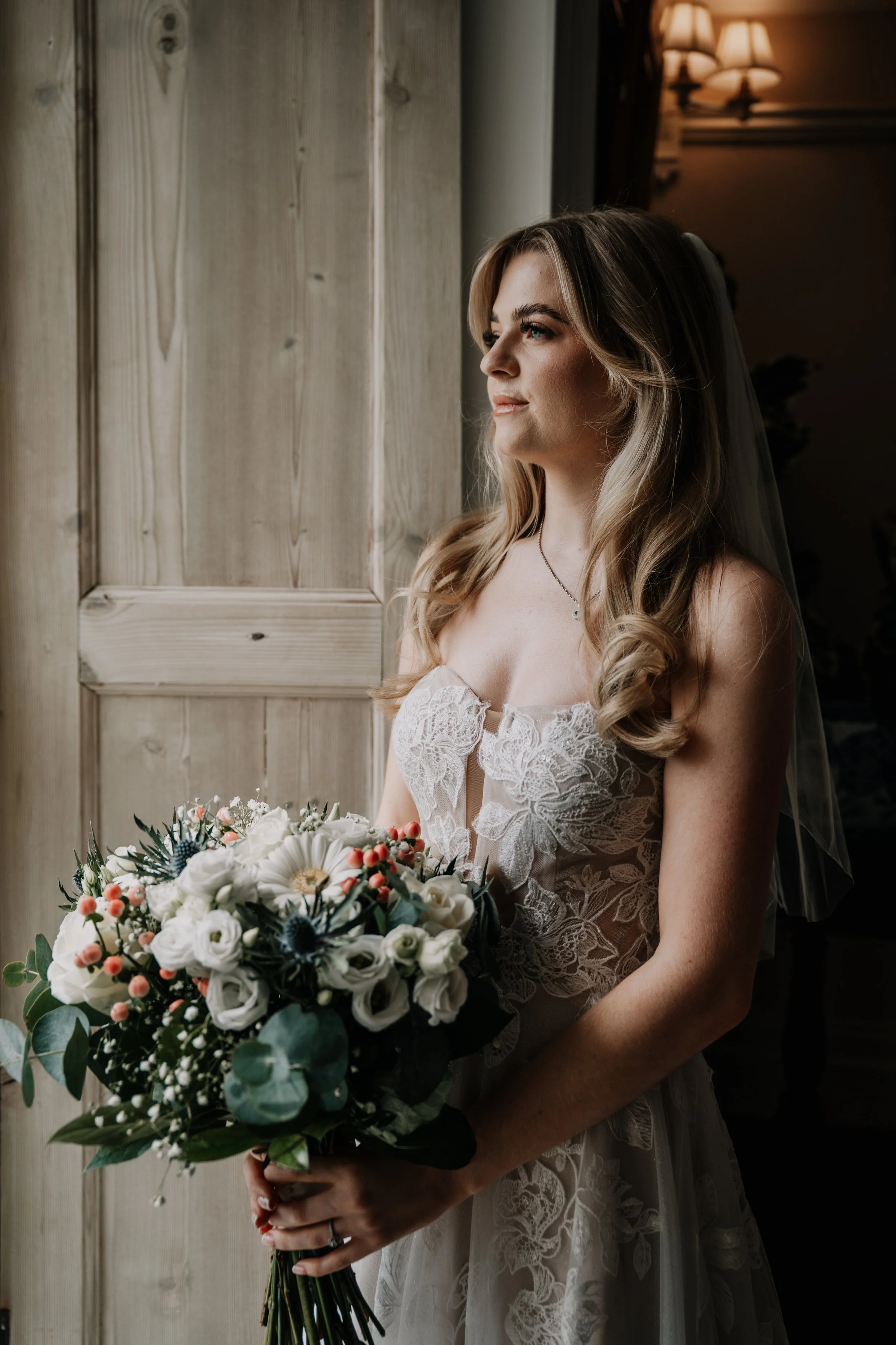 A bride holding a bouquet, standing indoors near a wooden wall, looking out a window.