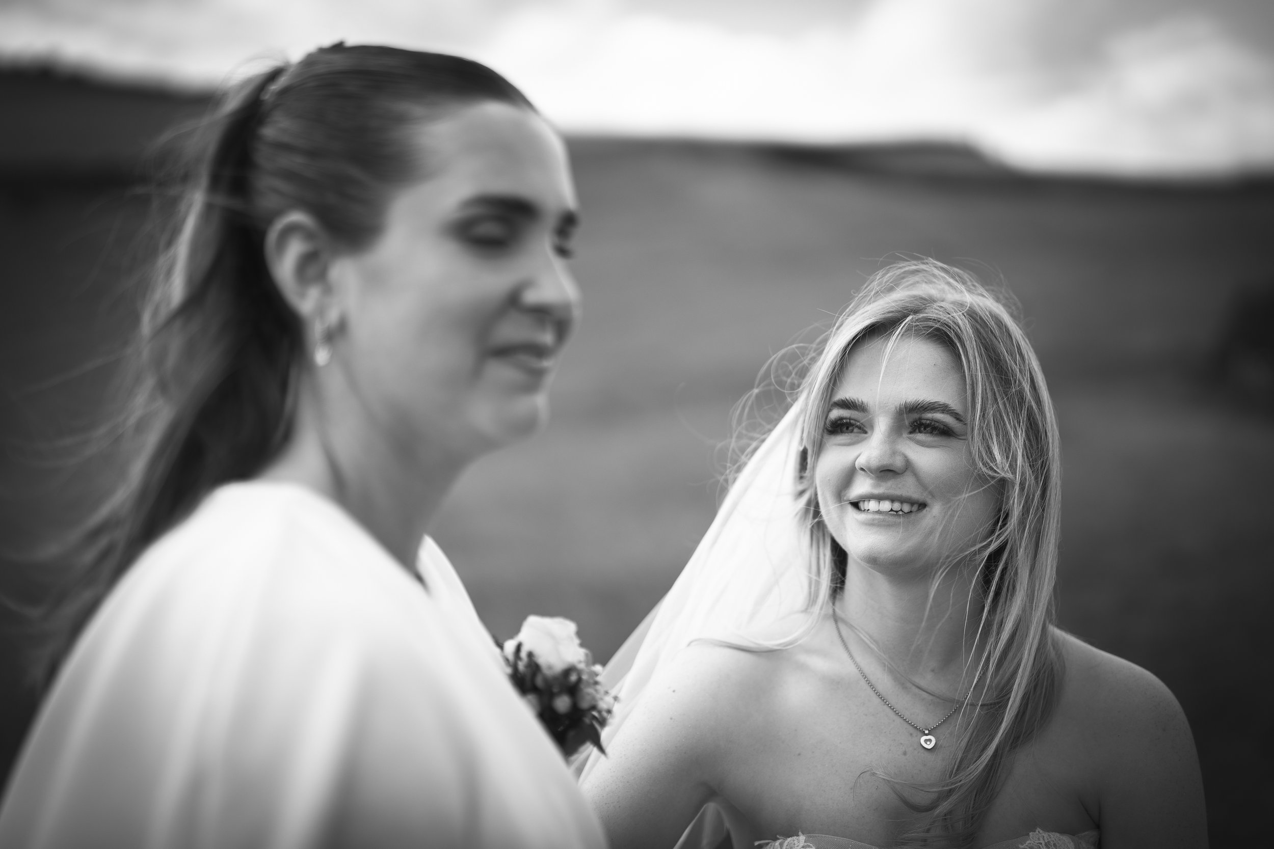 Black and white photo of two women outdoors, with one woman smiling at the other, who appears to be a bride with a veil and a flower bouquet on her chest, against a scenic landscape background.
