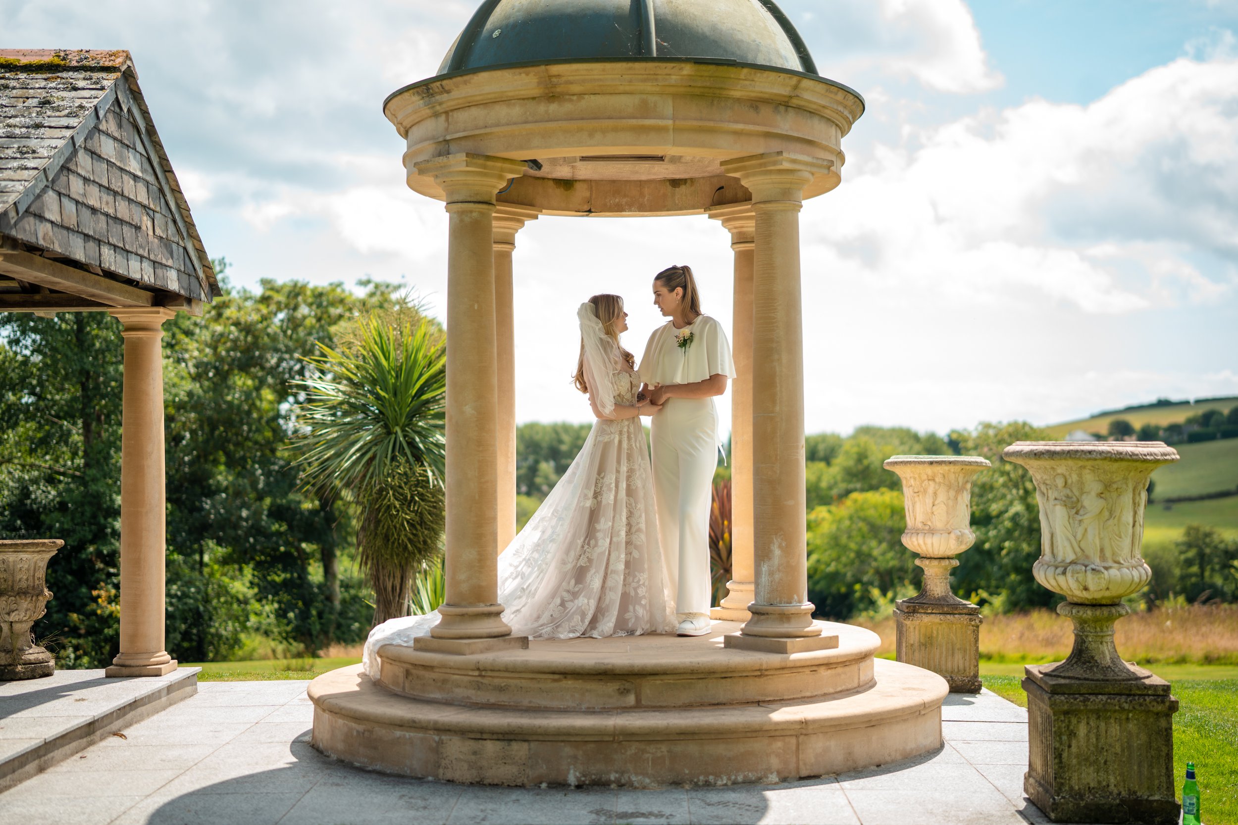 A bride and groom standing together in a stone gazebo outdoors, holding hands, with a scenic green landscape in the background on a sunny day.