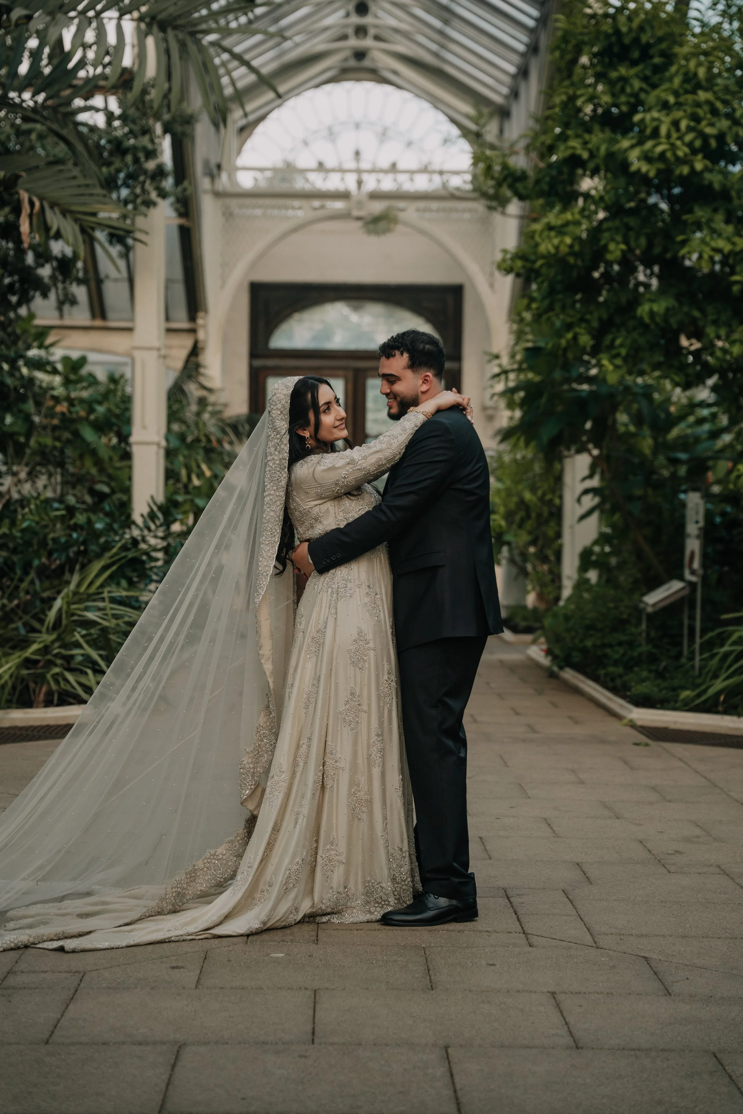 A bride and groom embrace in an indoor garden with lush greenery and a glass ceiling.