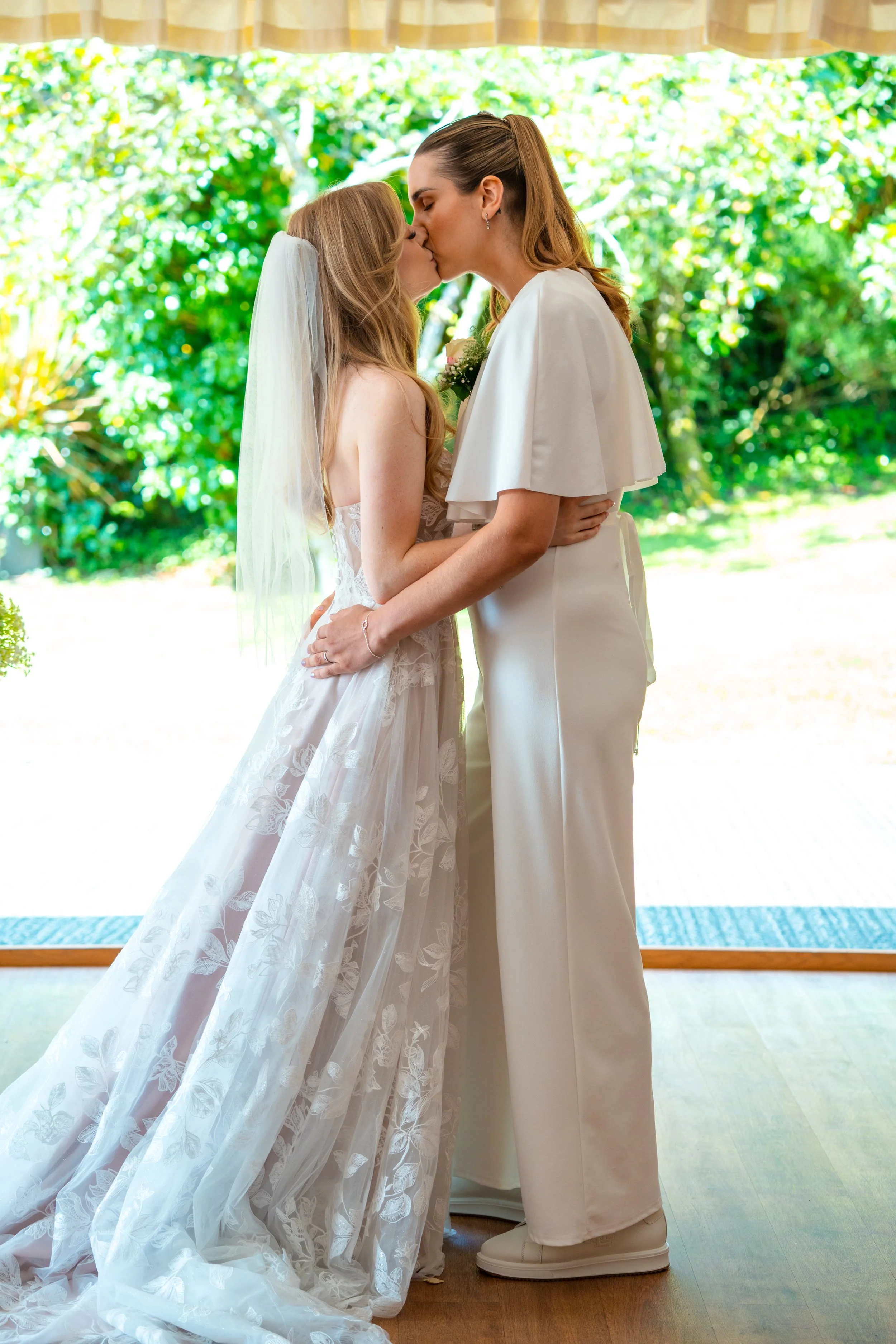Two women, likely at a wedding, sharing a kiss indoors with a window showing a garden background. One is in a wedding dress and veil, the other in a white outfit.
