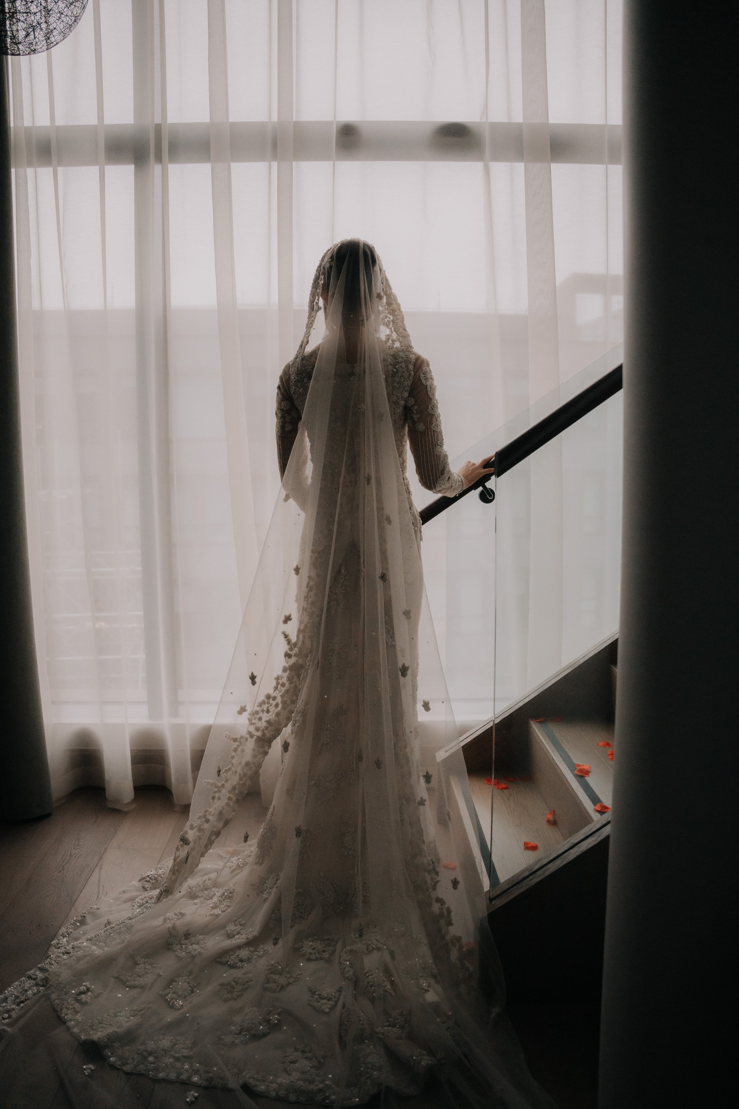 A bride in a wedding dress and veil standing on stairs by a large window, holding the railing with rose petals scattered on the step below.