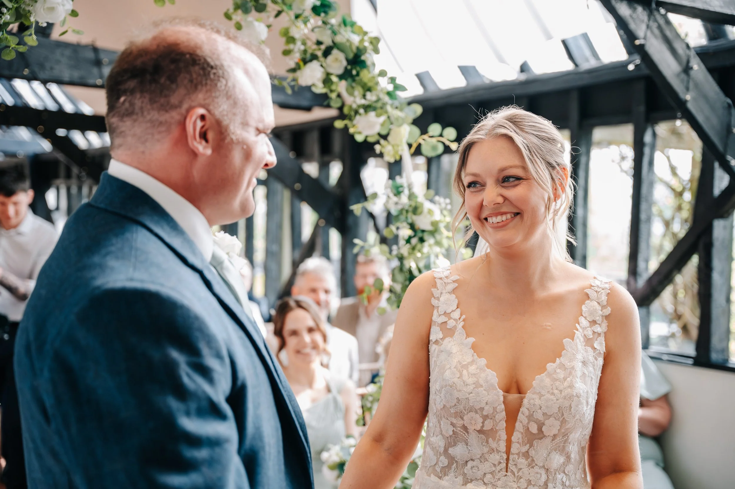 A bride and groom exchanging vows at their wedding ceremony, smiling at each other, with wedding guests seated behind them and floral decorations above.