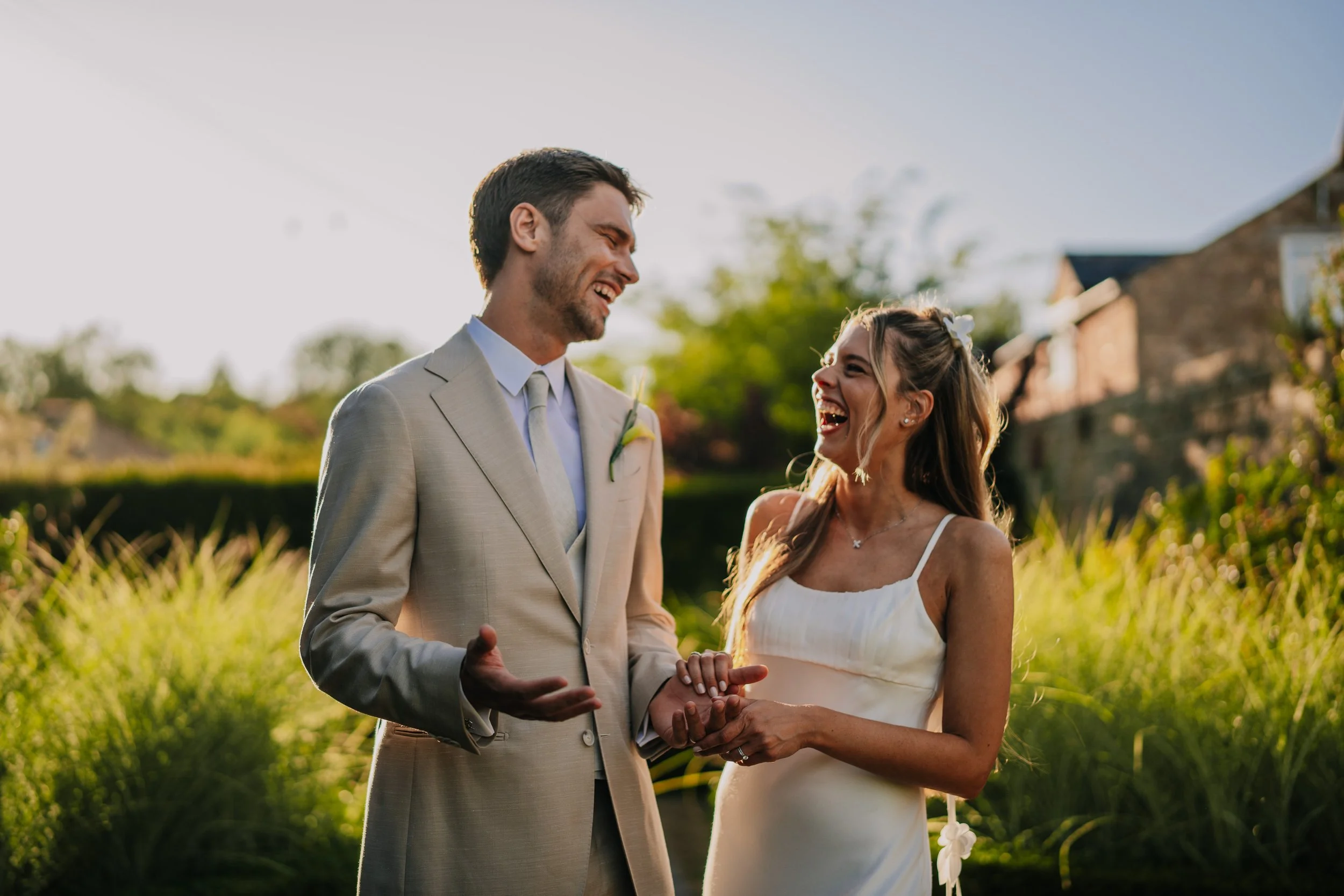 A couple dressed in wedding attire, holding hands and laughing outdoors during a sunny day.