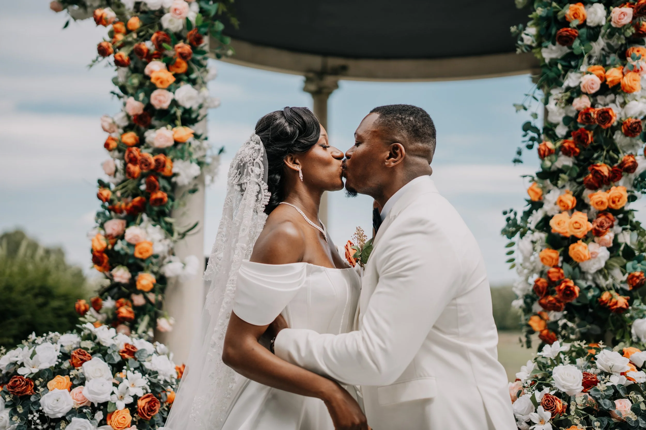 A bride and groom kiss during their outdoor wedding ceremony, framed by a floral arch with orange, white, and pink roses, against a partly cloudy sky.
