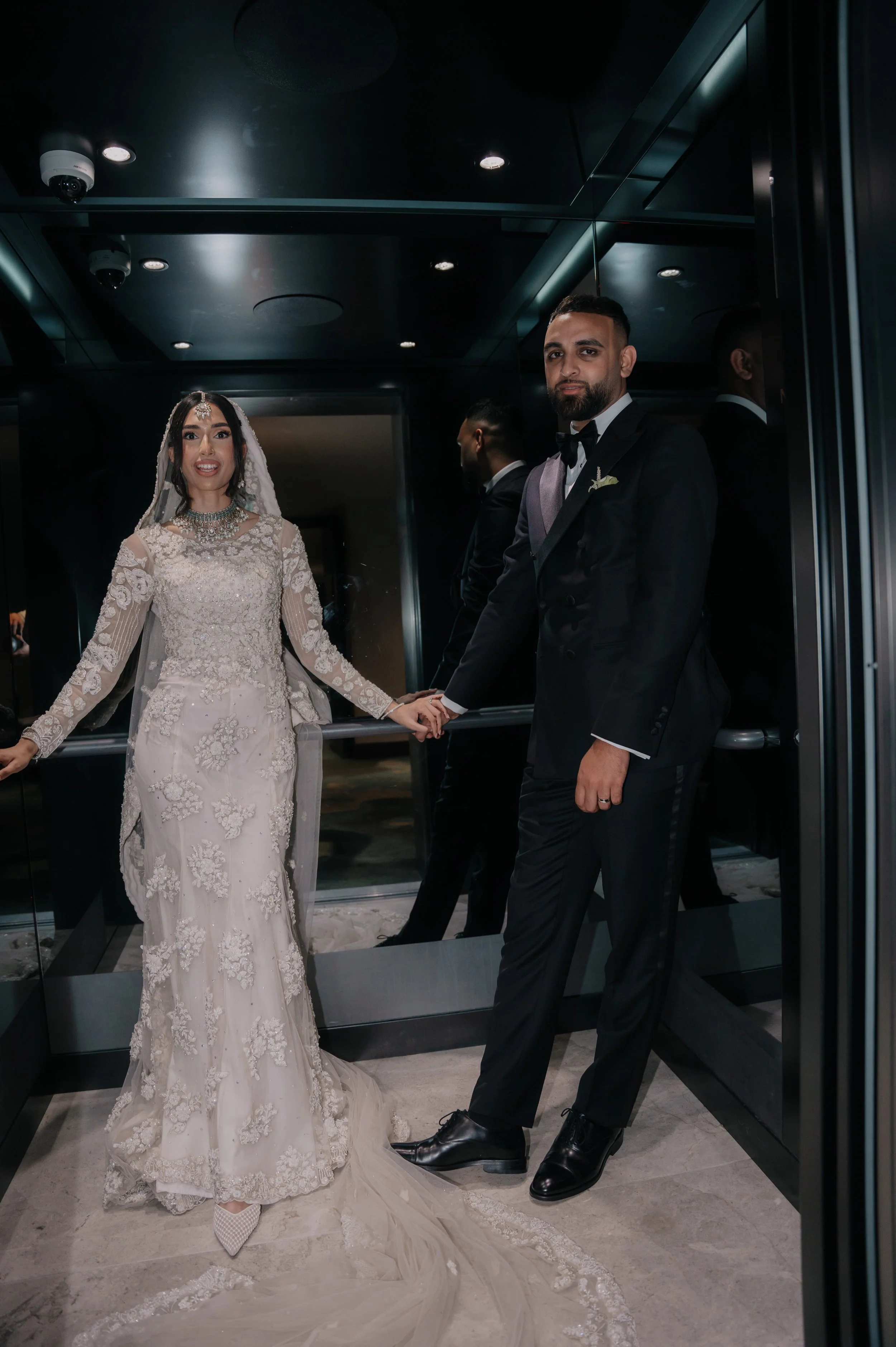 Bride and groom holding hands inside an elevator, dressed in wedding attire, with the bride in a white lace gown and the groom in a black tuxedo.
