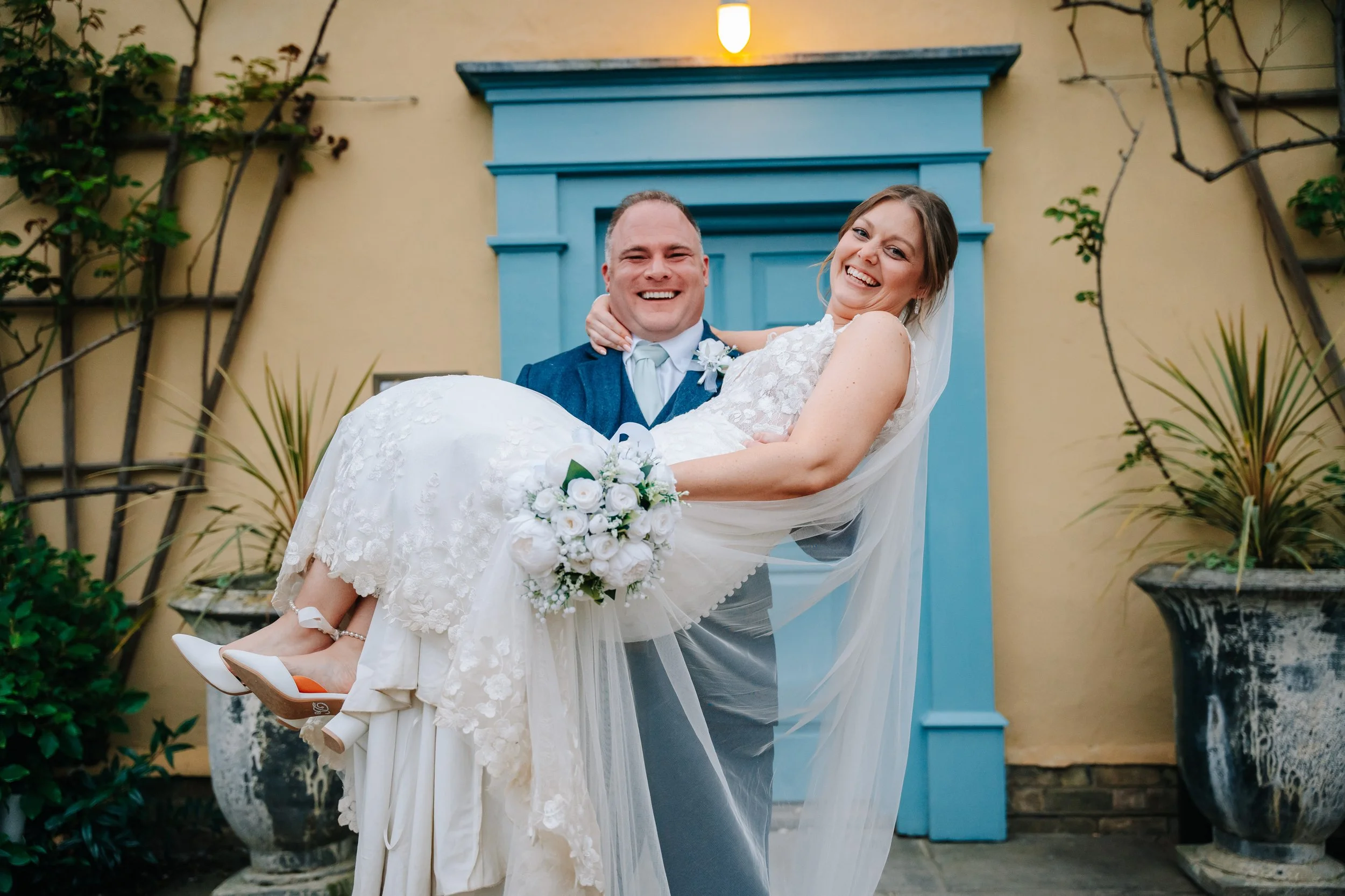 A groom lifting a smiling bride holding a bouquet of white flowers outdoors, with decorative planters and a yellow wall with a blue window frame in the background.