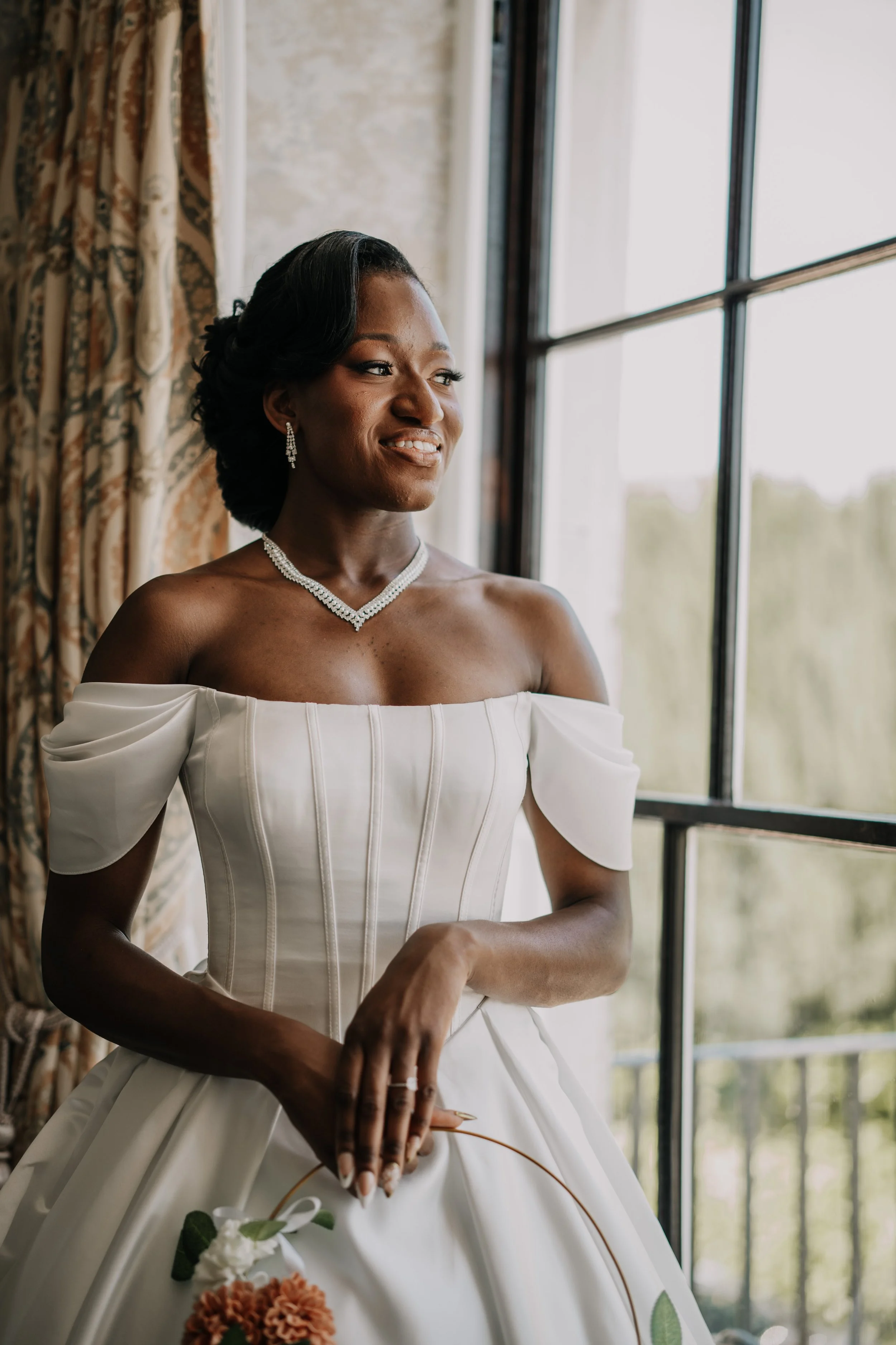 A woman in a white off-the-shoulder wedding dress with puffed sleeves, standing by a window with a scenic view outside. She is wearing jewelry, including earrings and a necklace, and holding a floral bouquet.