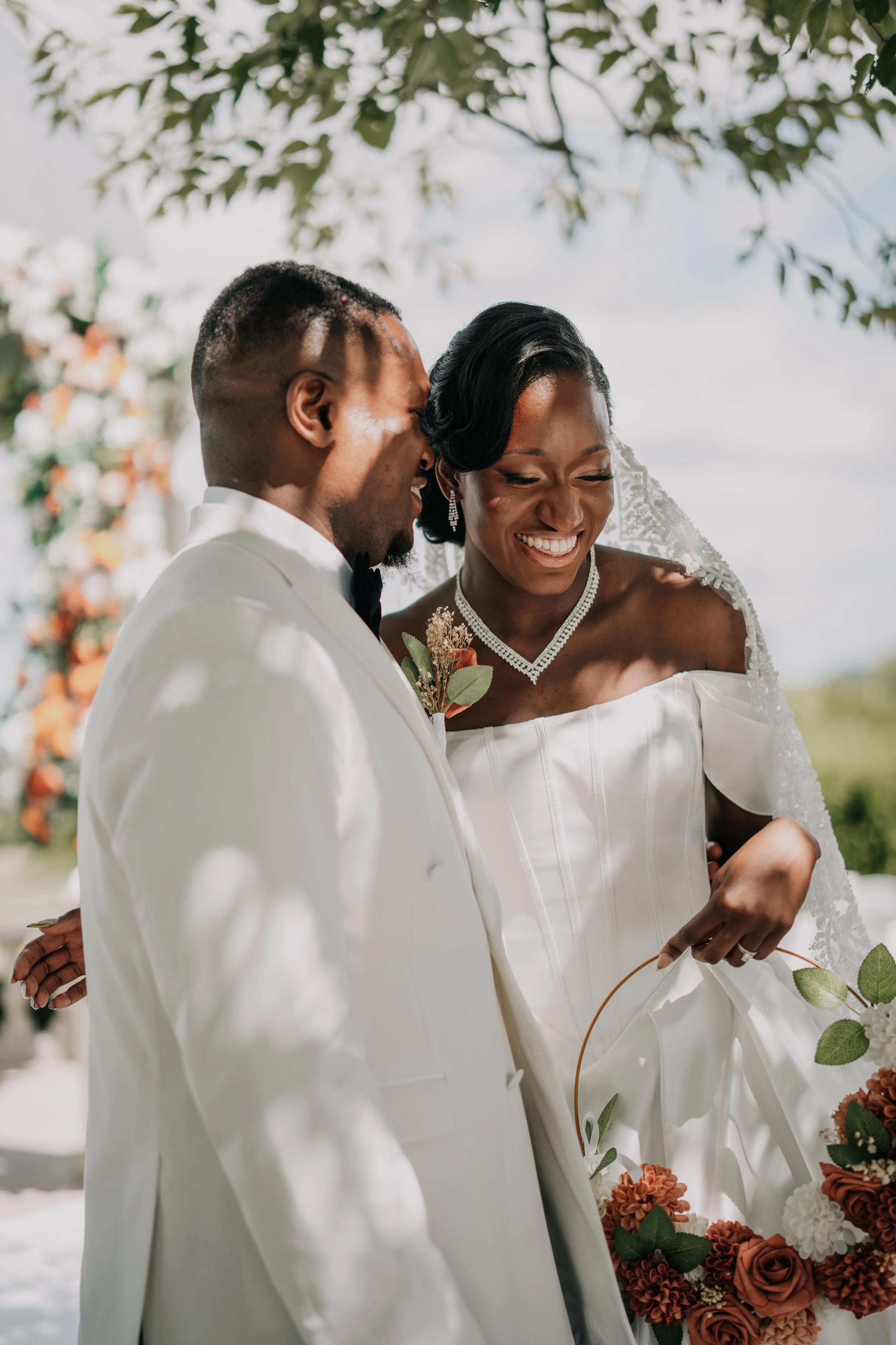A bride and groom sharing a joyful moment at their outdoor wedding, standing close with genuine smiles, with floral decorations and greenery in the background.