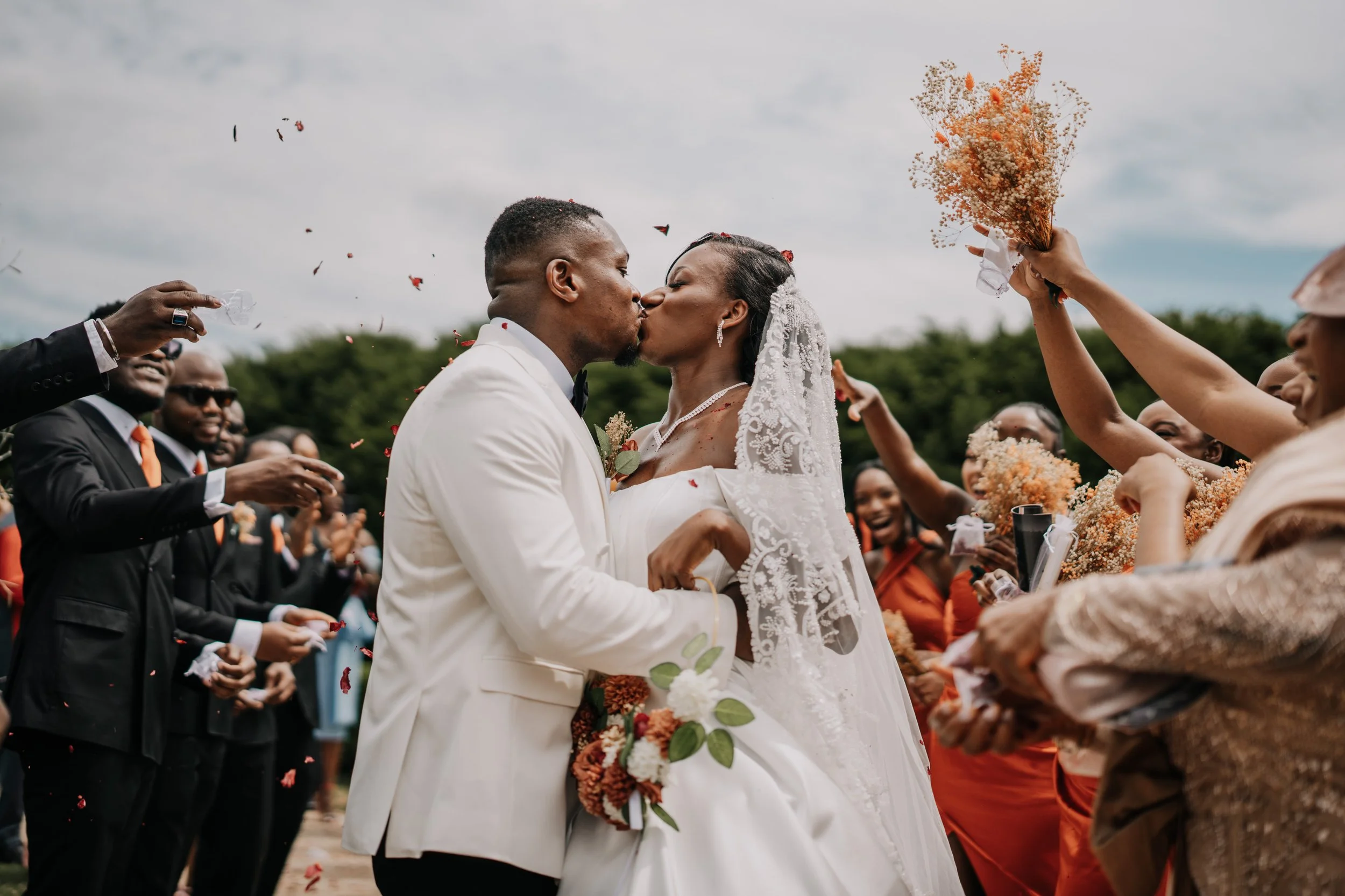 A newlywed couple sharing a kiss during their outdoor wedding ceremony, surrounded by friends and family throwing flower petals and holding bouquets under a cloudy sky.