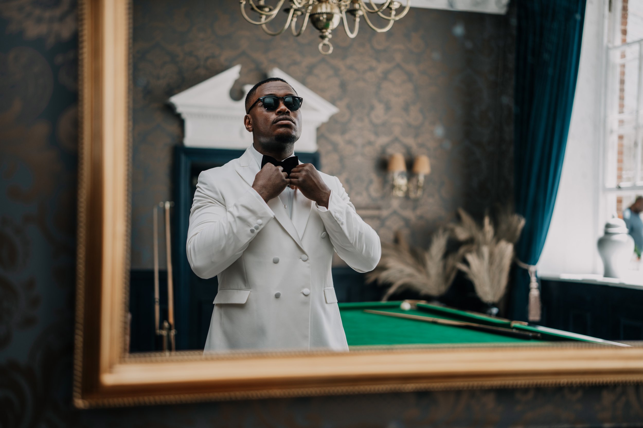 A man in a white tuxedo adjusting his black bow tie in front of a mirror, wearing sunglasses in a stylish room with a pool table, golden wall decor, and blue curtains.