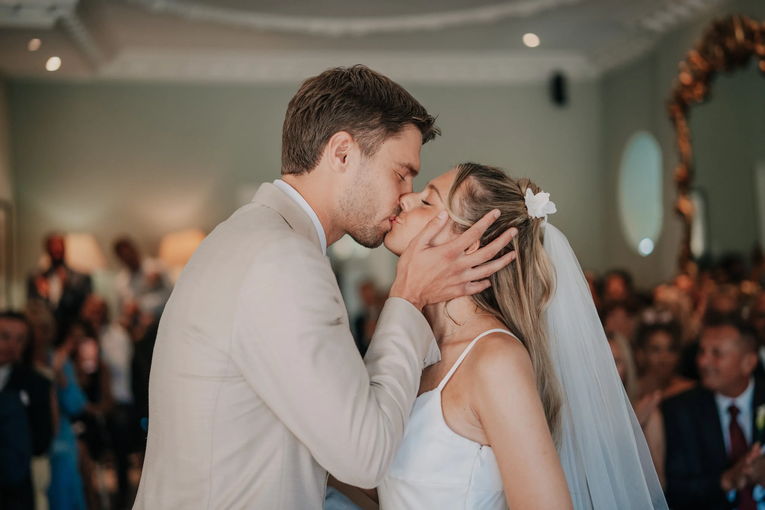 A bride and groom share a kiss during their wedding ceremony, with the groom holding the bride's face and the bride wearing a white dress and veil in a decorated indoor setting.