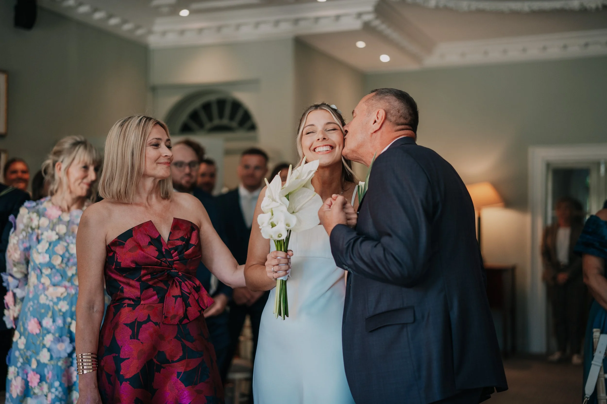 A bride in a white dress holds a bouquet of white calla lilies and smiles as her father kisses her cheek during a wedding ceremony, with guests watching in the background.