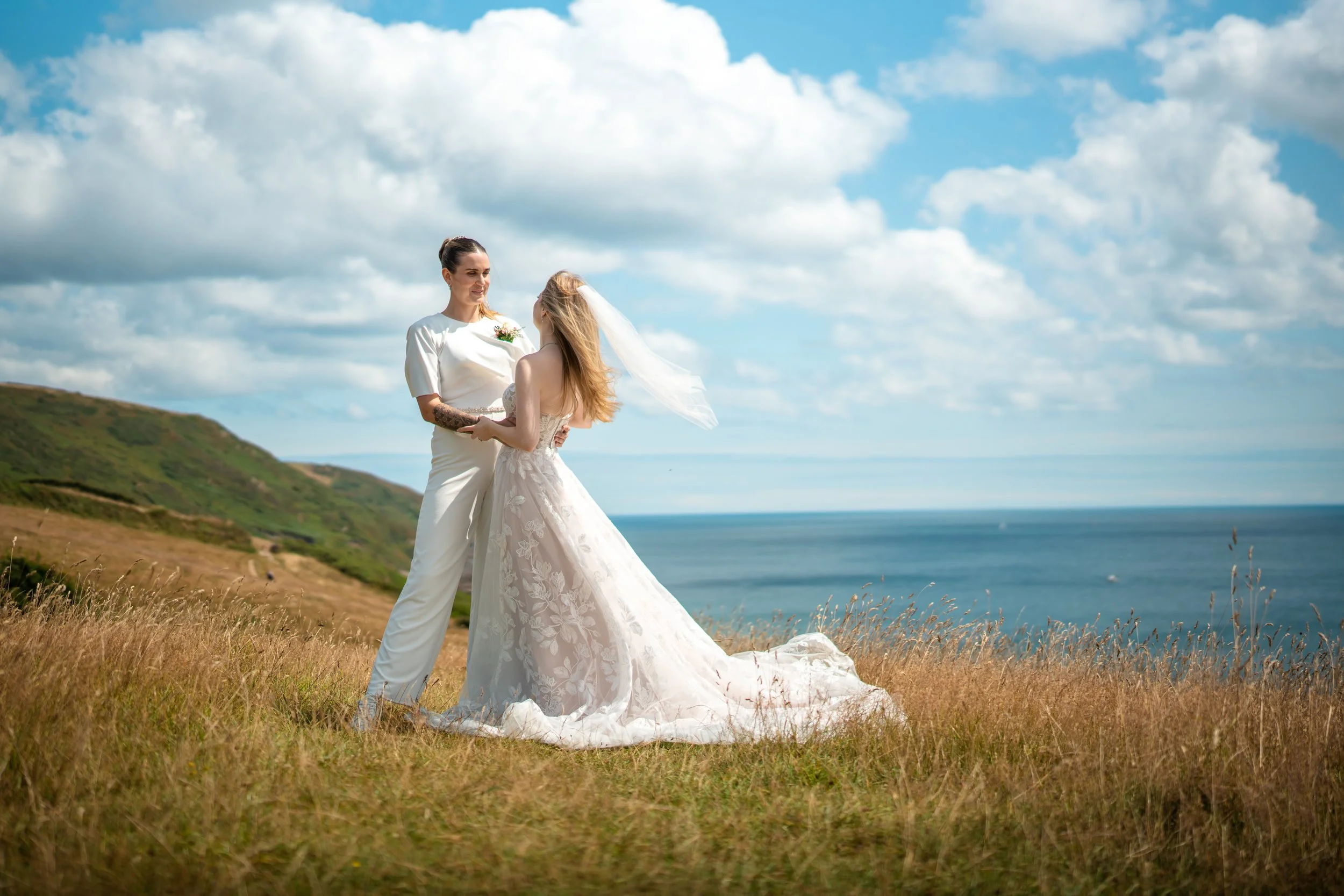 Two women in wedding dresses stand outdoors on a grassy hillside with the ocean and a cloudy sky in the background.