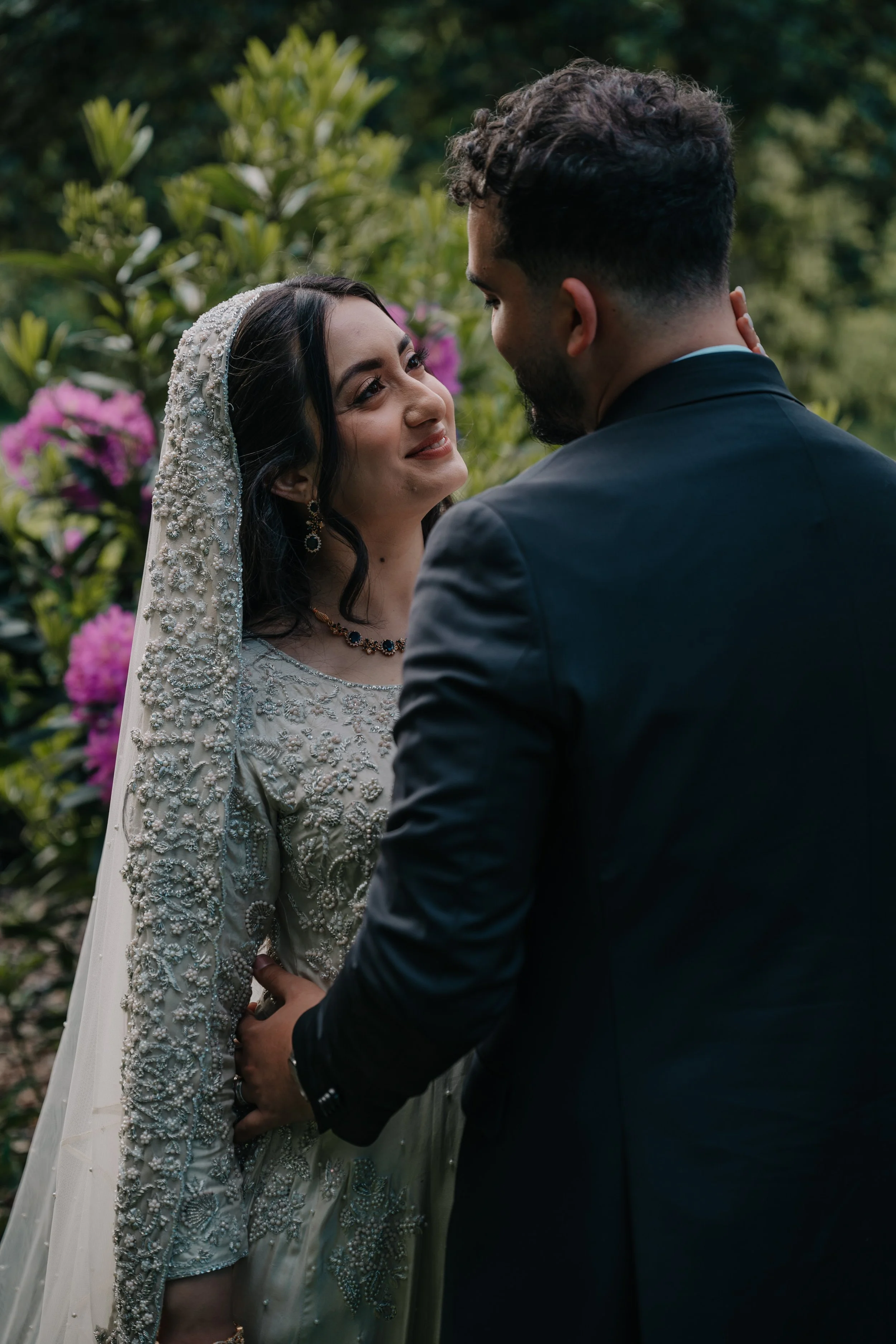 A bride and groom are standing outdoors, gazing into each other's eyes with a background of green foliage and pink flowers.