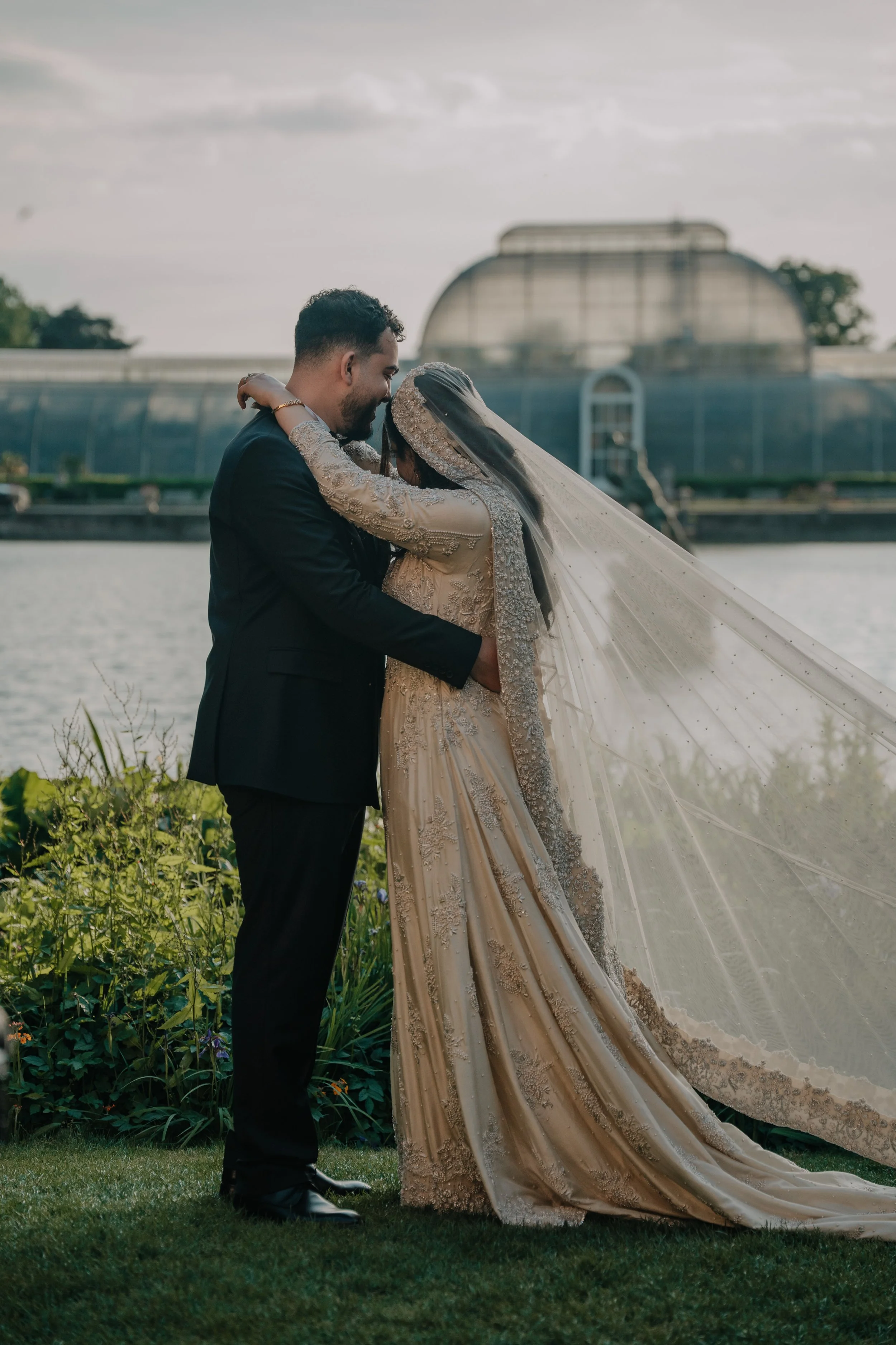 A bride and groom sharing a kiss outdoors by a lake, with a greenhouse in the background. The bride is wearing a beige embroidered wedding dress and veil, and the groom is in a black suit.