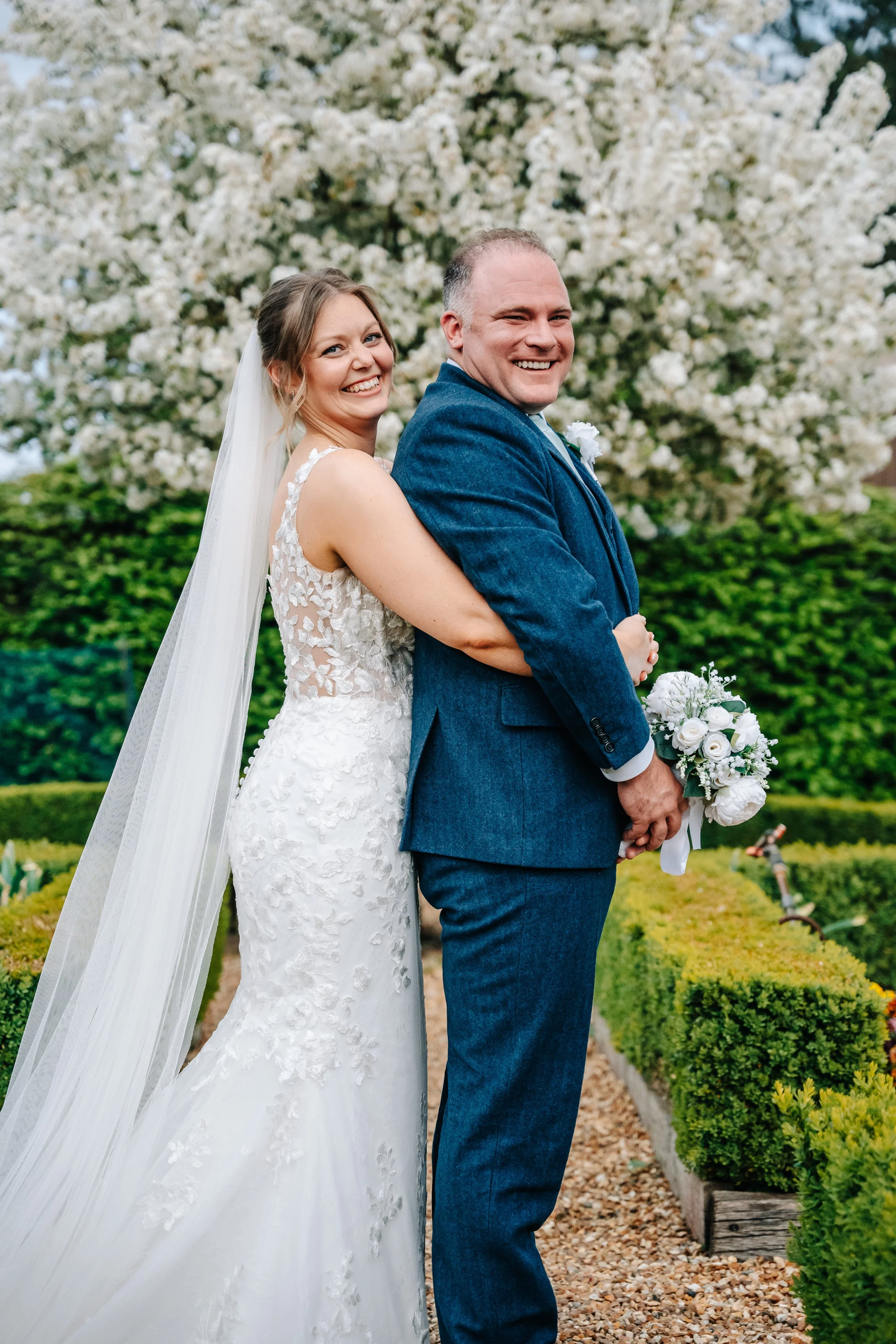 A bride and groom in wedding attire smiling and standing back-to-back in a garden with blooming trees and green bushes.
