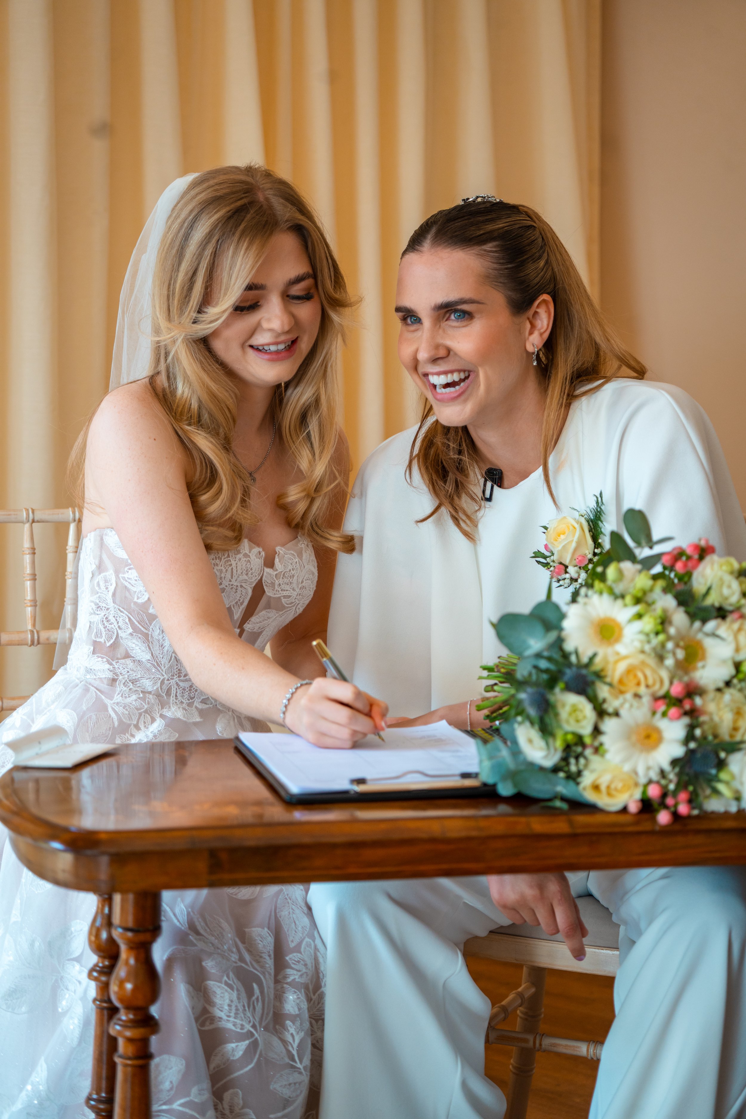 Two women, one in a wedding dress and the other in white, sit at a wooden table signing documents during a wedding ceremony, with a bouquet of flowers nearby.