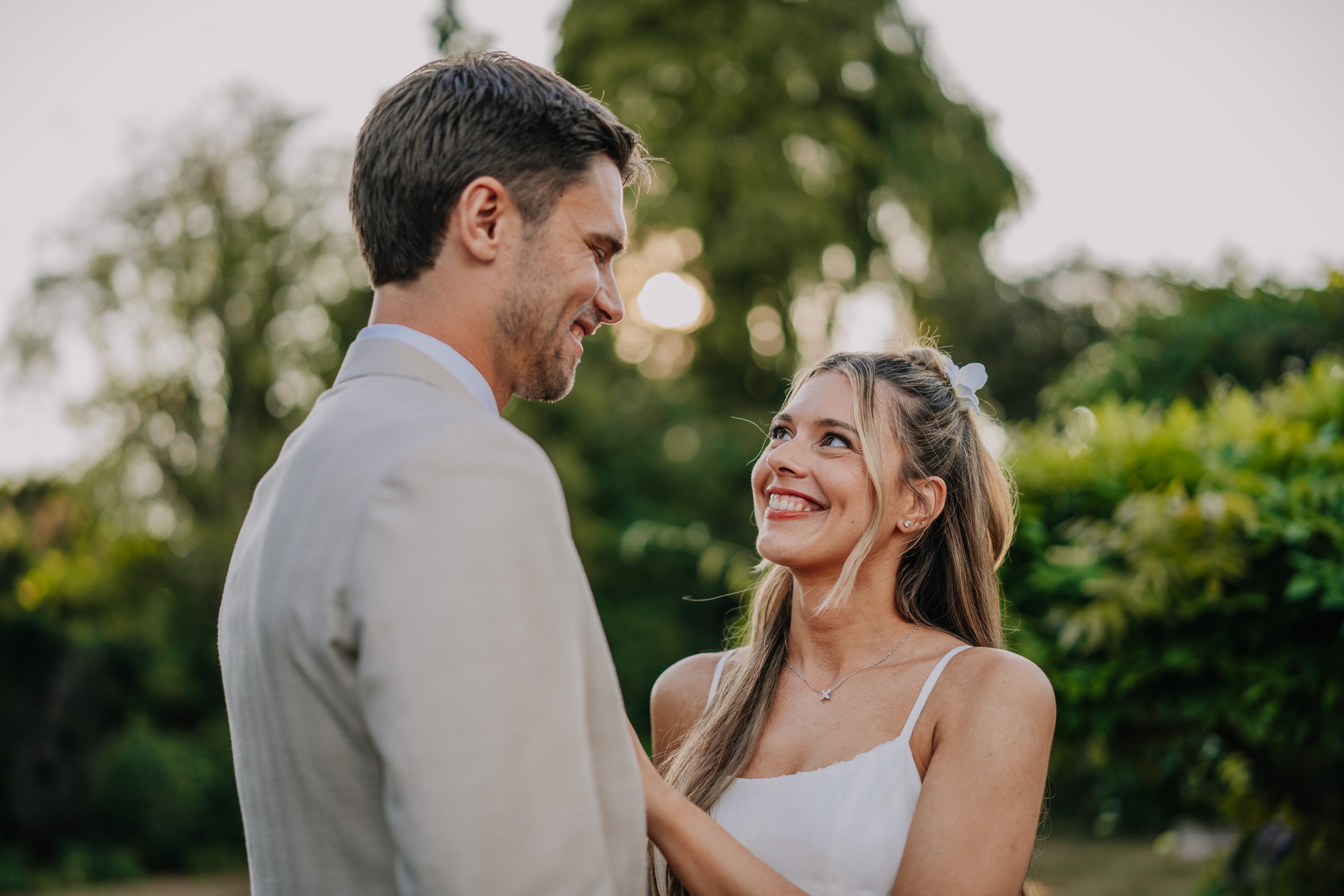 A couple, dressed in wedding attire, smiling at each other outdoors surrounded by greenery.