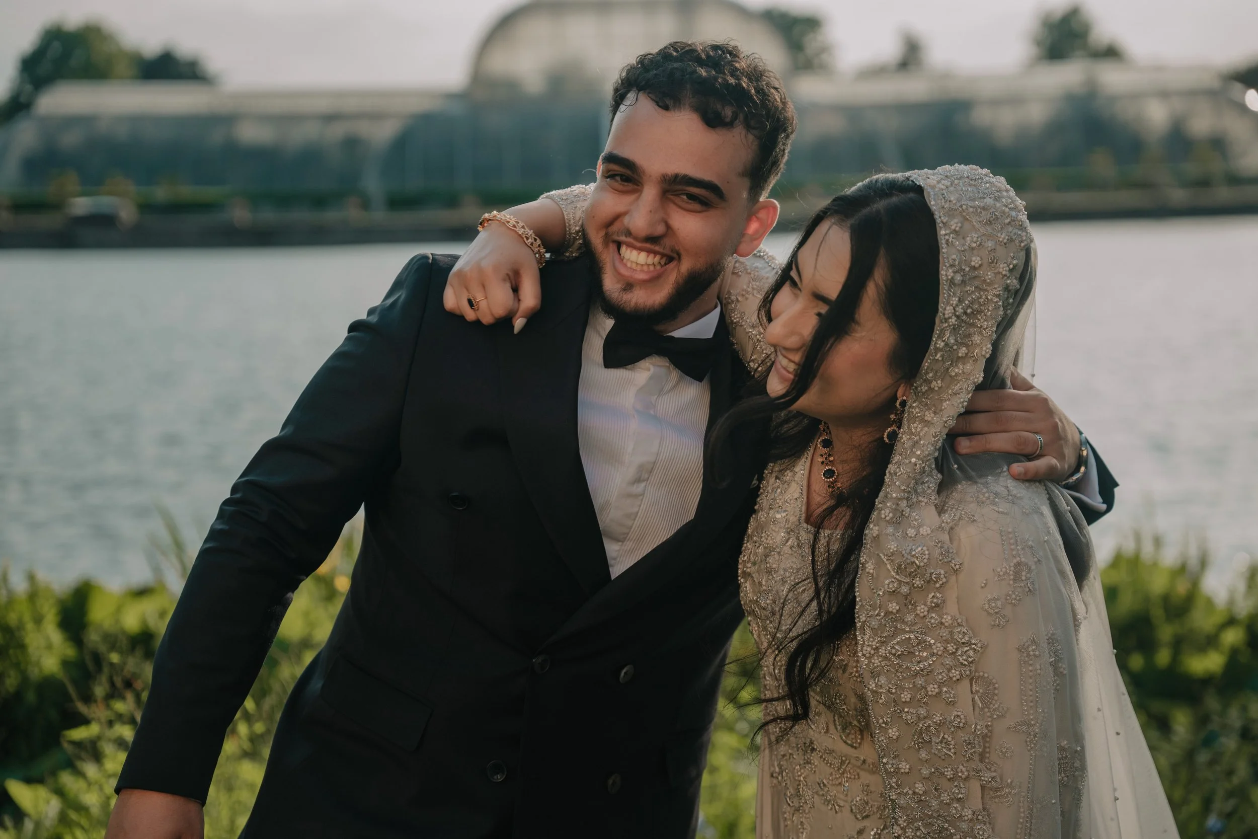 A newlywed couple smiling and embracing each other outdoors by a river, with a bridge and green landscape in the background.