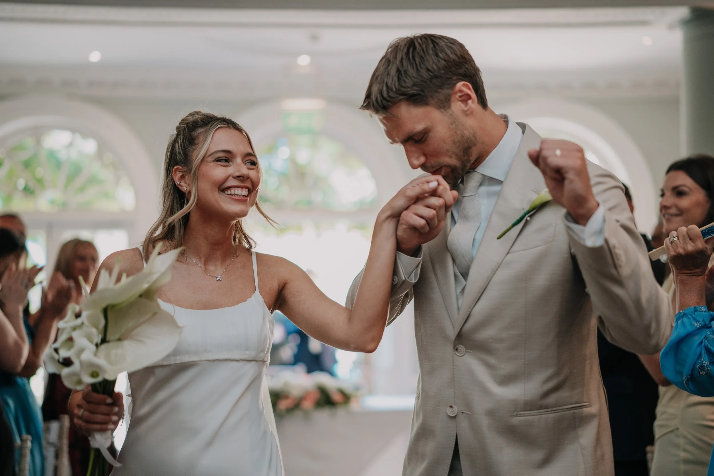 A bride holding a bouquet of white calla lilies smiling as she kisses her groom on the hand during their wedding ceremony indoors with guests in the background.