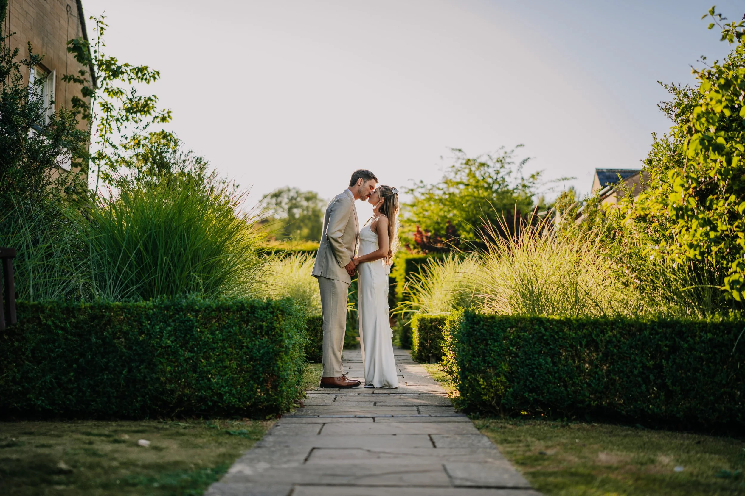 A couple dressed in wedding attire standing close together on a stone path in a garden, their foreheads touching and holding hands, with green bushes and trees in the background and sunlight shining.
