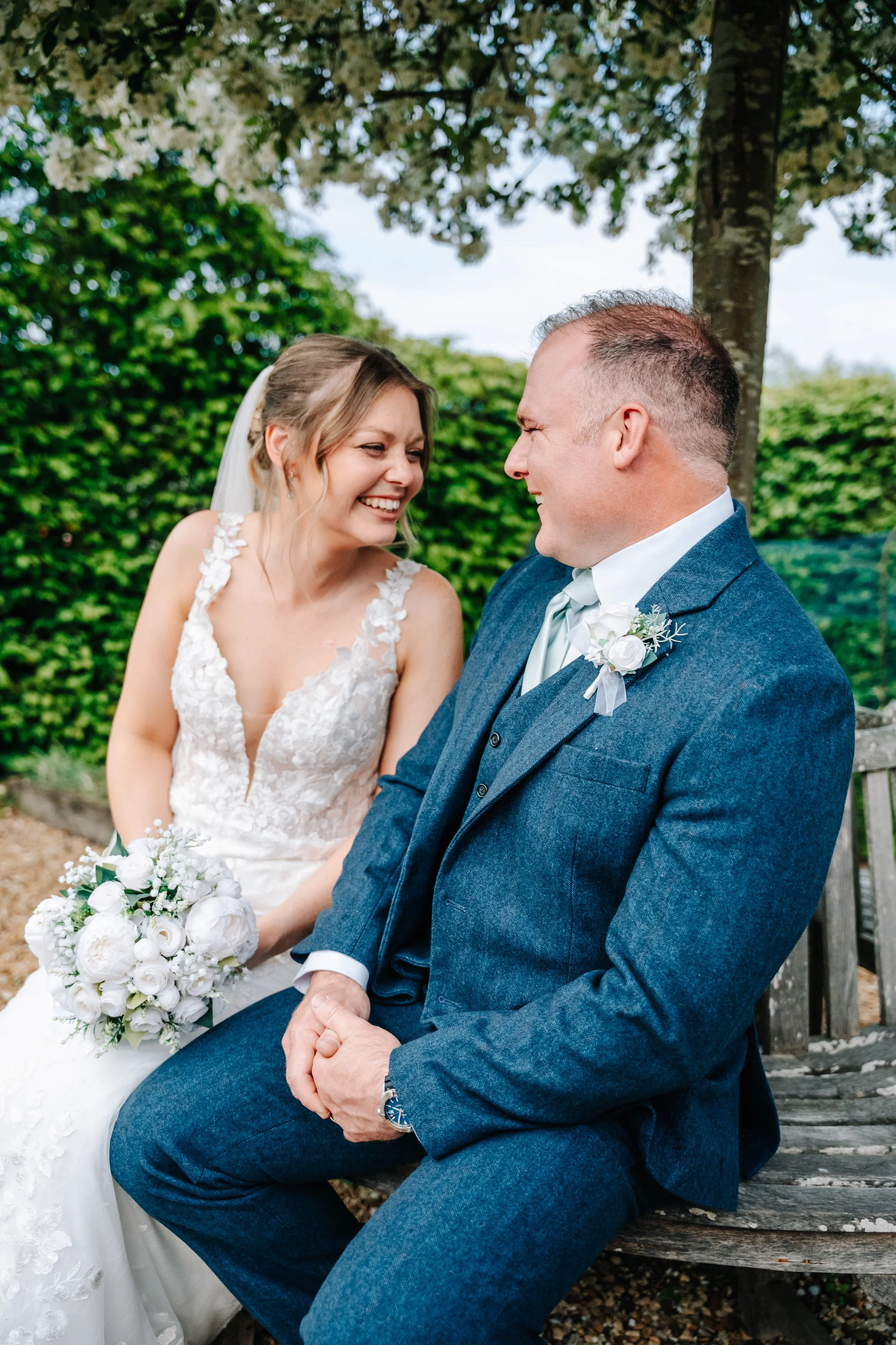 A bride and groom sitting on a wooden bench outdoors, smiling and looking at each other, surrounded by green foliage.