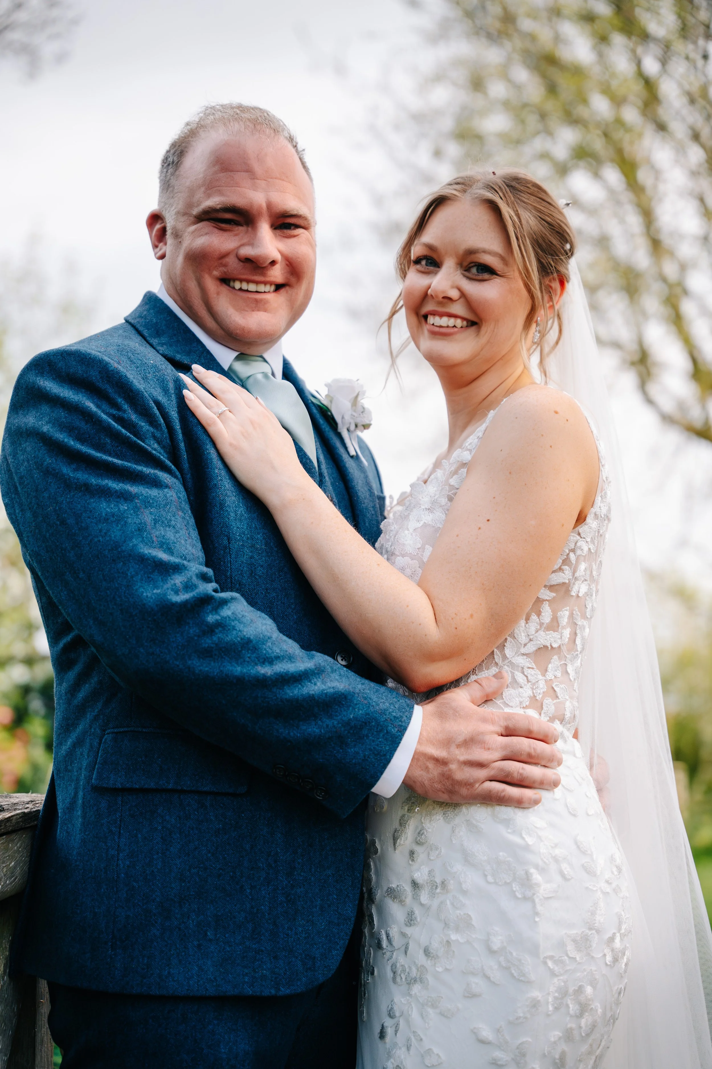 A wedding couple smiling outdoors, the man in a blue suit and light green tie, and the woman in a white lace wedding dress with a veil.