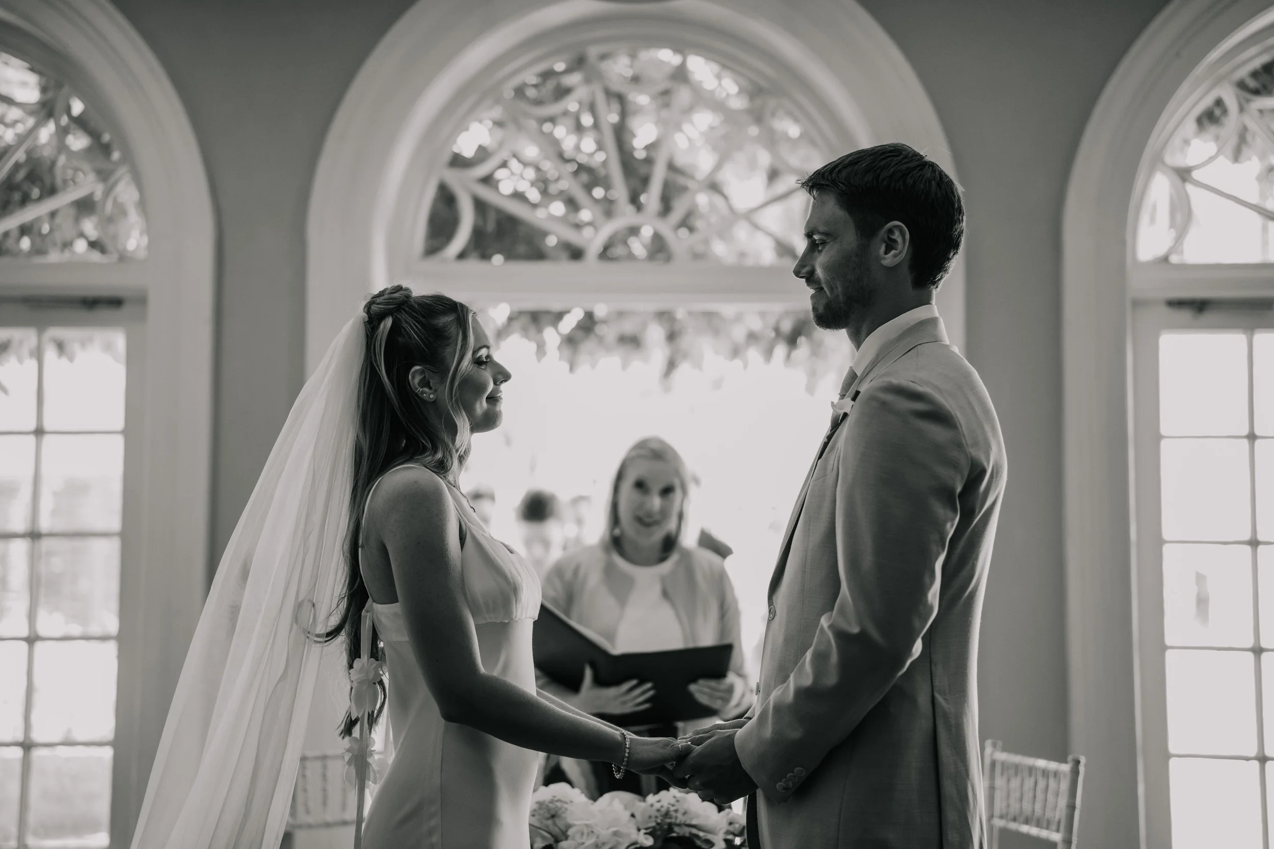 A black and white photo of a bride and groom holding hands during their wedding ceremony in a room with large windows and ornate arches, with an officiant standing behind them.