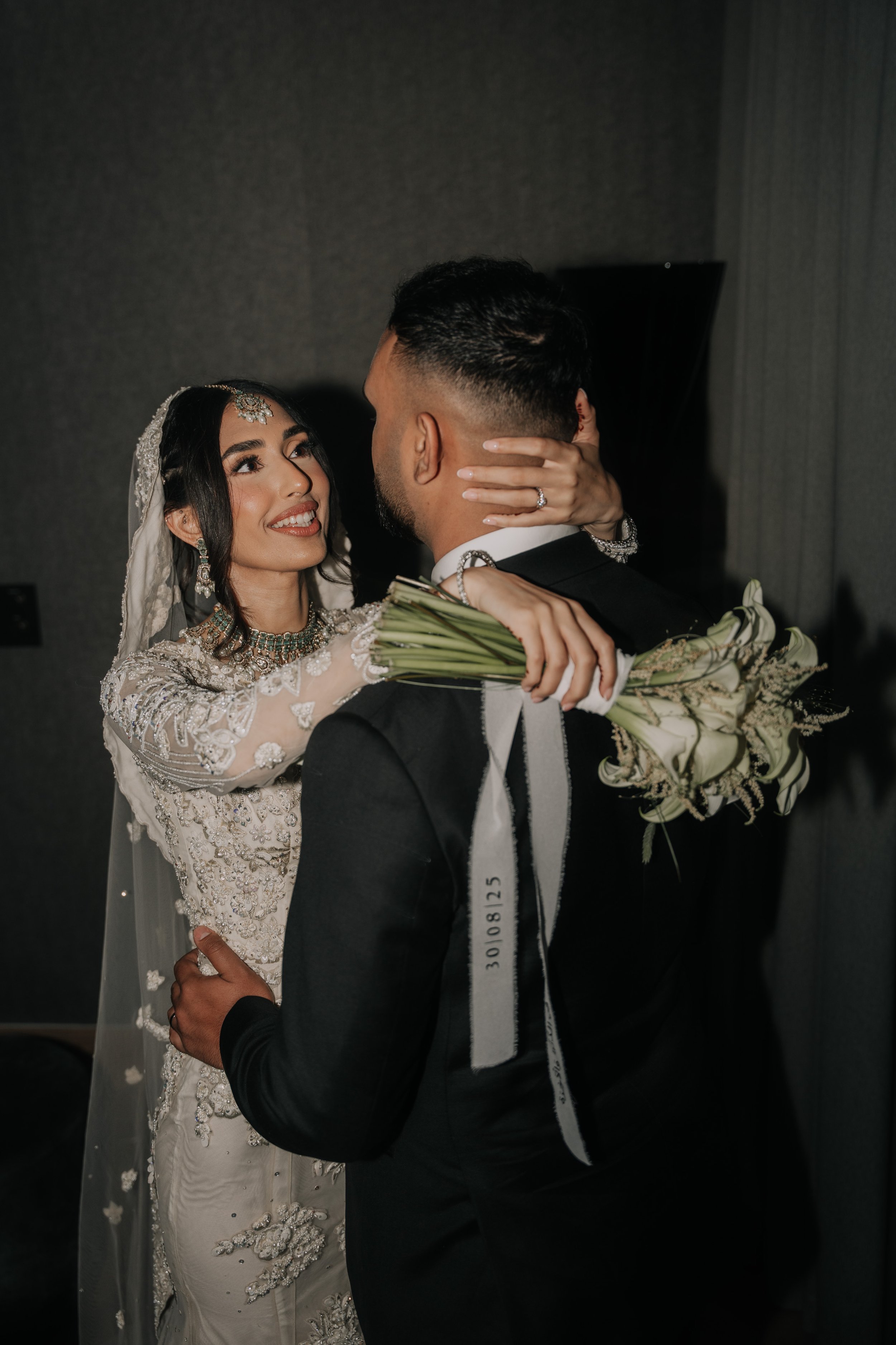 A bride and groom embrace on their wedding day, with the bride holding a bouquet of flowers and wearing a detailed wedding dress and jewelry, while the groom wears a black suit.