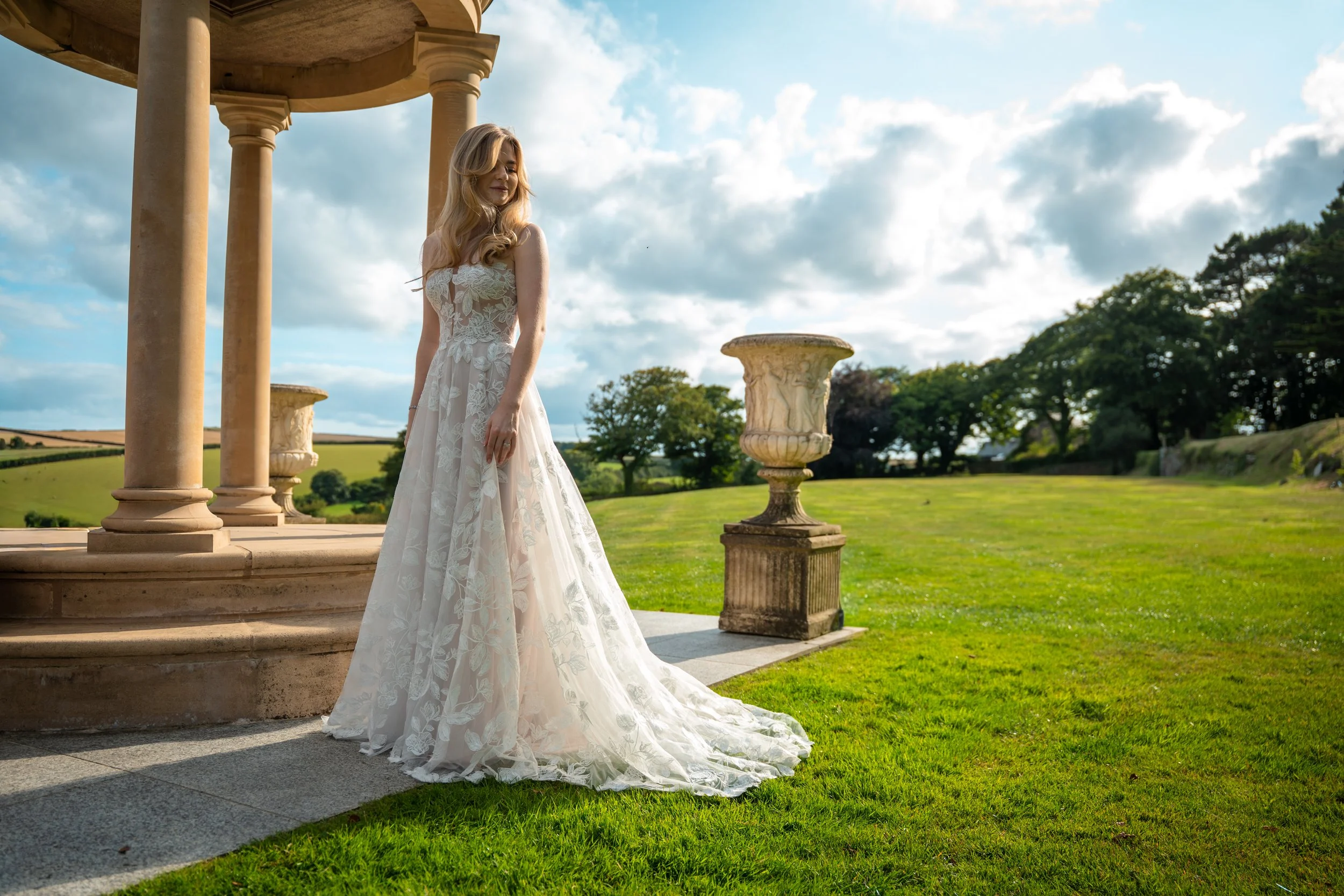 A woman in a white lace wedding dress standing outside near a stone structure with columns, with green grass and trees under a partly cloudy sky.