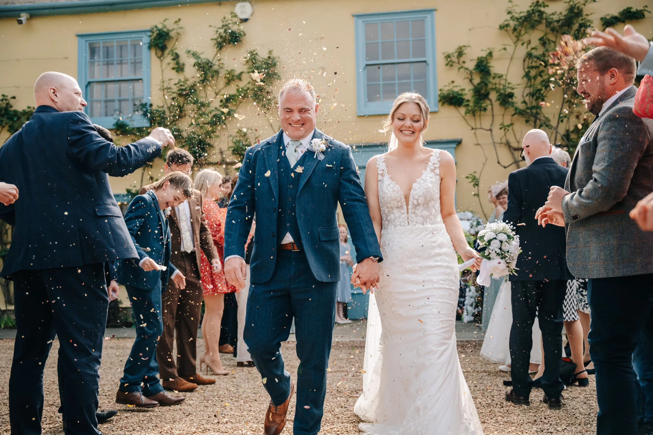 The newly married bride and groom walking hand in hand through a crowd of wedding guests throwing confetti outside a yellow house with blue window frames.
