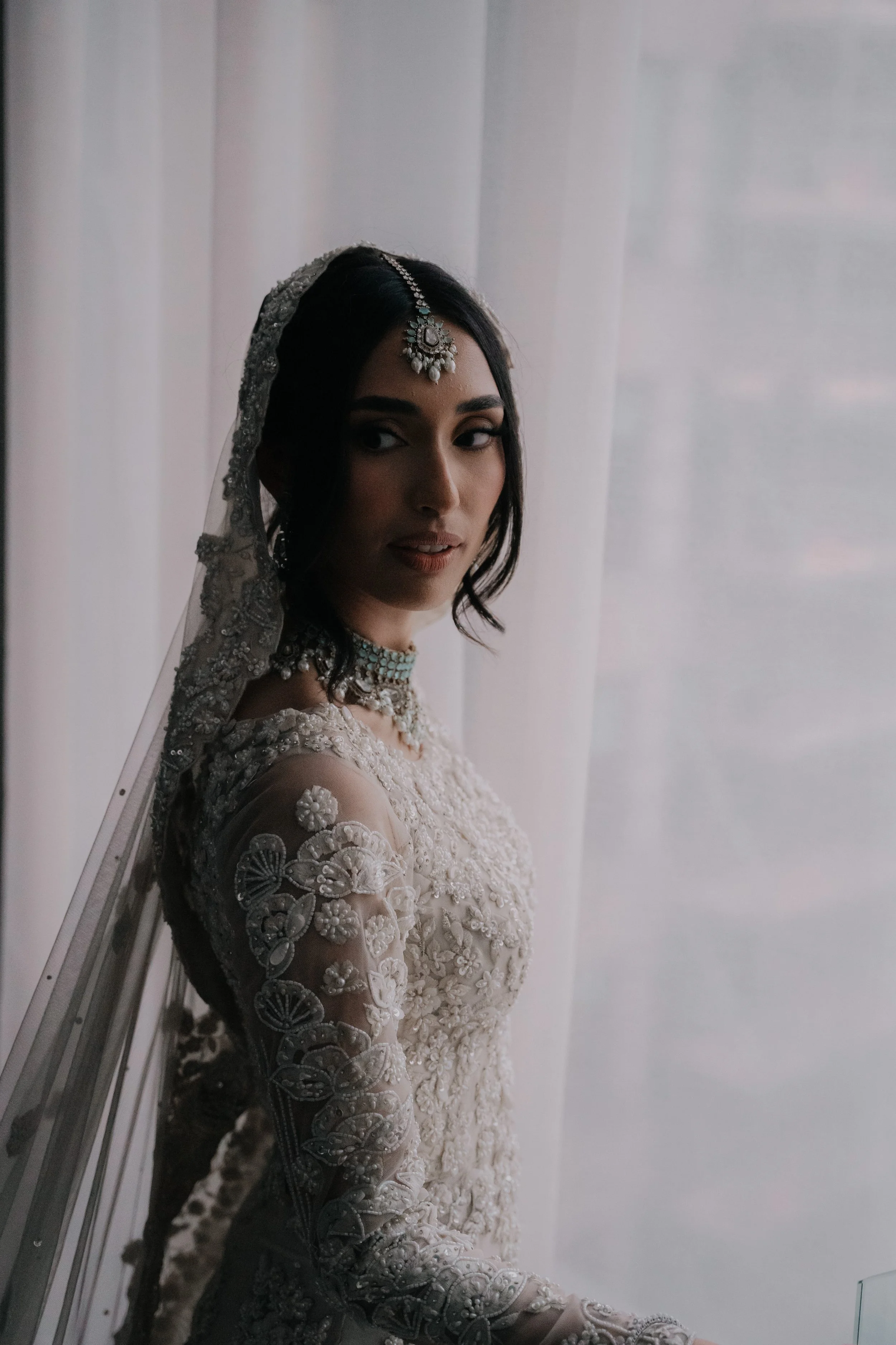 A woman in a wedding dress with intricate embroidery and lace detail, wearing traditional jewelry, and standing near a sheer curtain, looking over her shoulder.