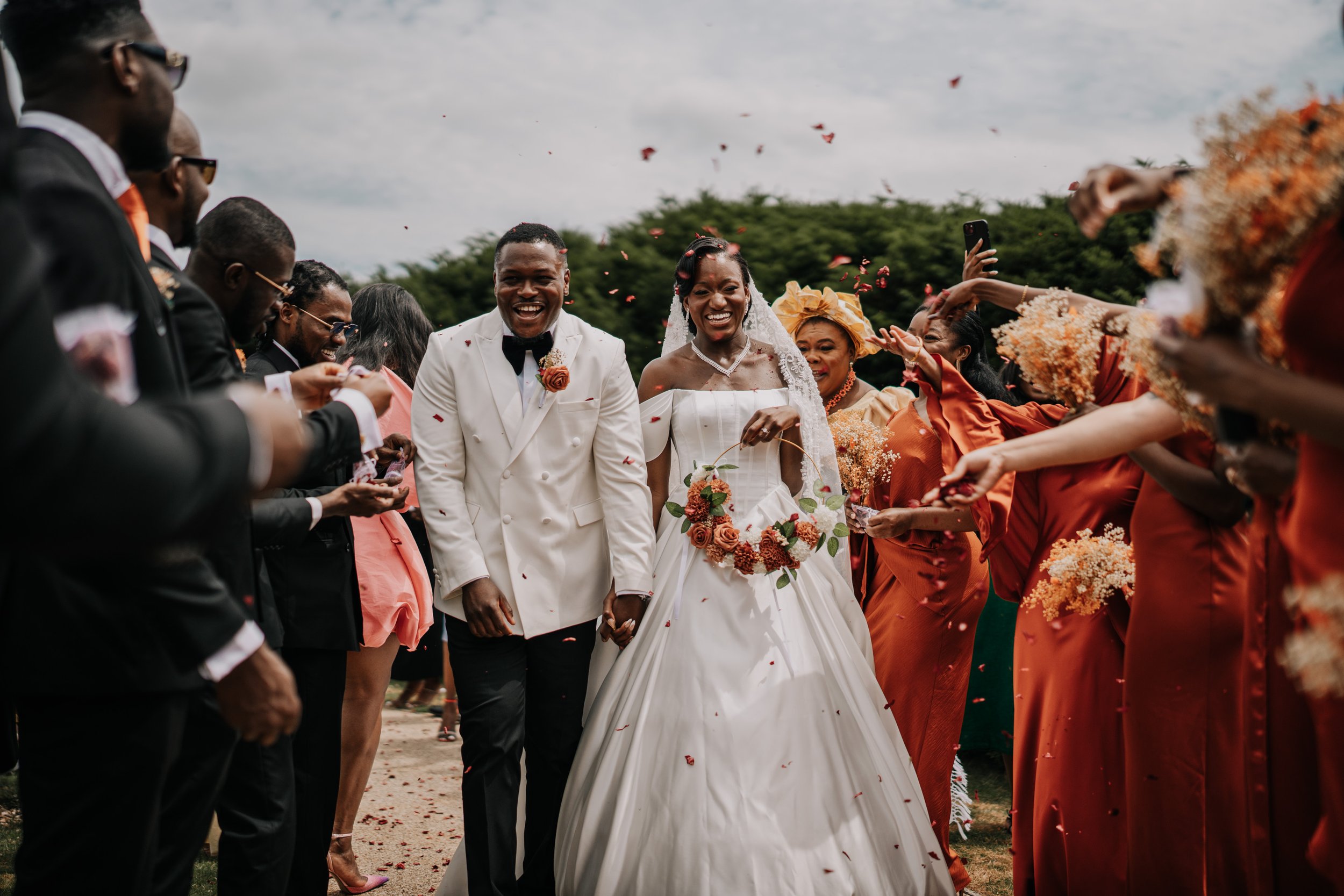 A bride and groom holding hands walking through a celebration procession of guests throwing petals, with guests dressed in black, pink, and orange dresses, outdoors on a cloudy day.
