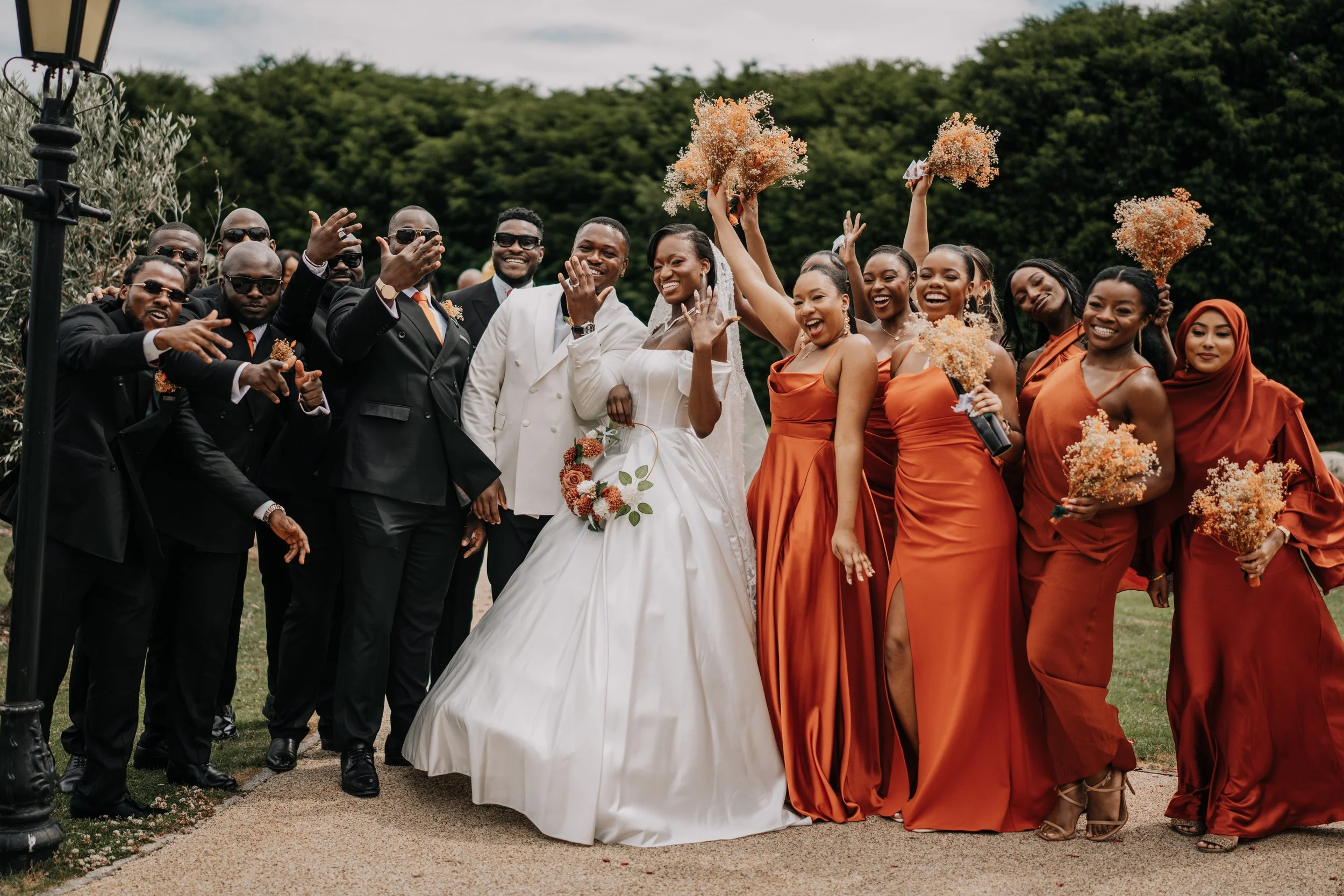 Group of groomsmen and bridesmaids celebrating at a wedding outdoors, with the bride and groom in the center. The bride is in a white wedding gown holding a bouquet, and the bridesmaids are in orange dresses holding bouquets. The groomsmen are in bla