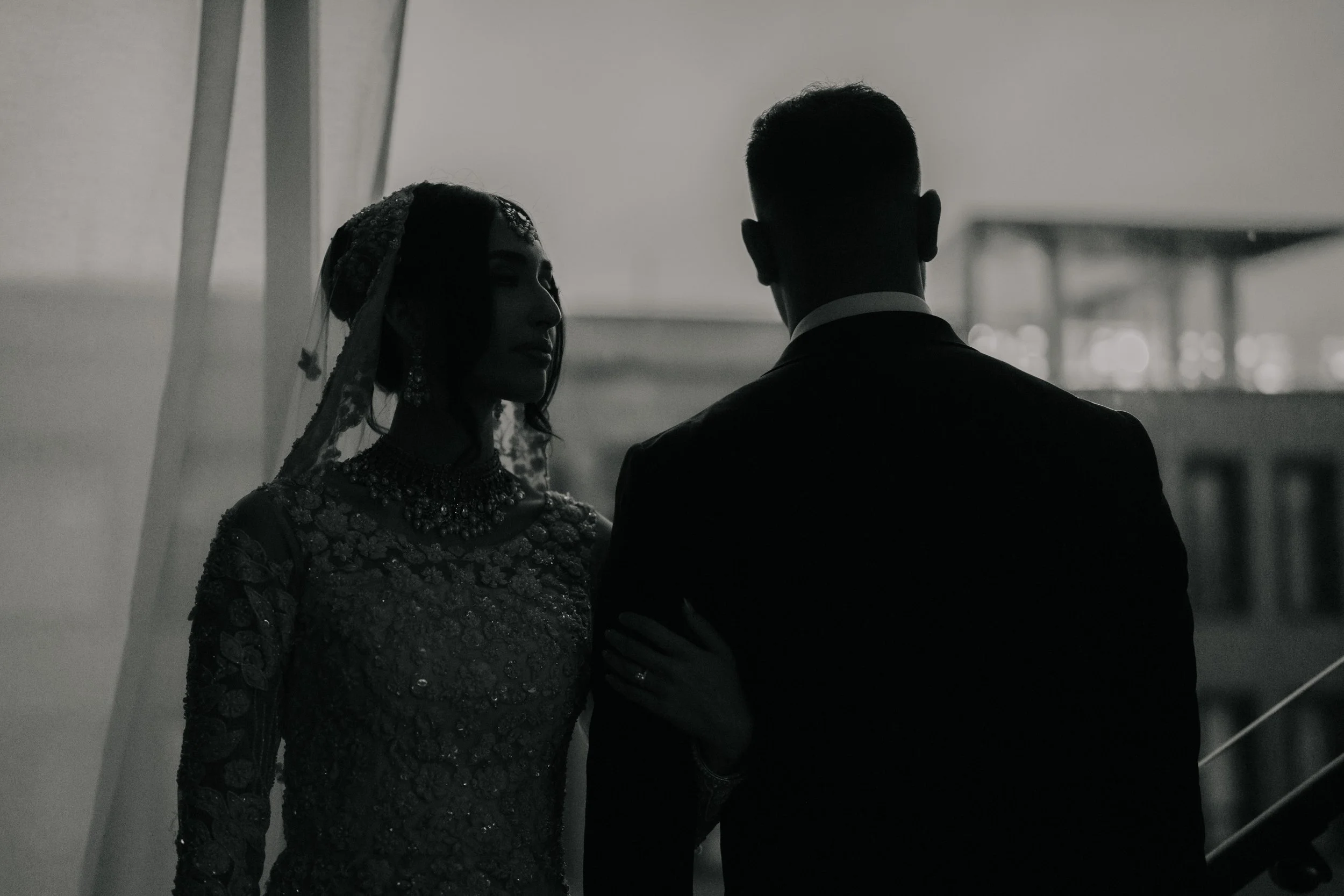 Black and white photo of a bride and groom standing close, with the bride examining the groom's arm, indoors with blurred background.